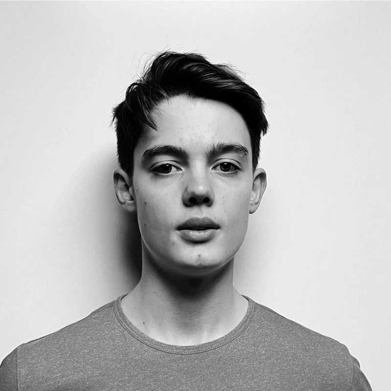 Black and white portrait of a young man with short, dark hair and a neutral expression, wearing a plain t-shirt, standing against a light background.
