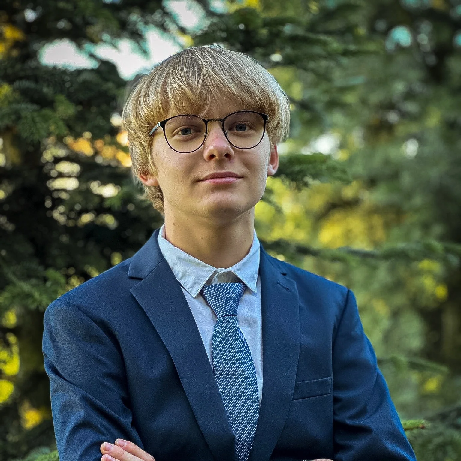 A young man with blond hair and glasses wearing a blue suit and tie, standing outdoors with trees in the background.