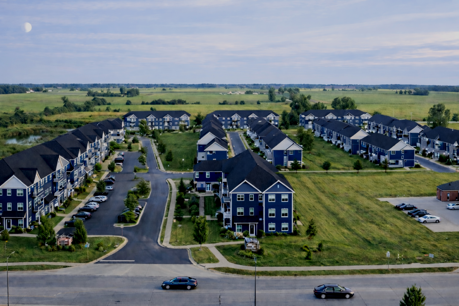 Aerial view of a residential apartment complex with multiple blue and white buildings, parking lots, and green lawns surrounded by open fields under a partly cloudy sky.