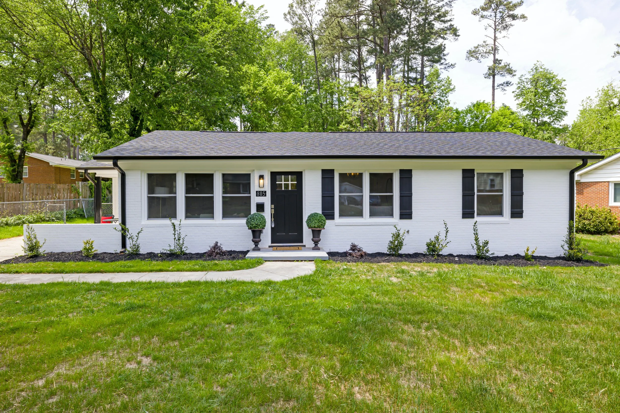 White single-story house with black shutters, front lawn, landscaped garden, surrounded by trees.