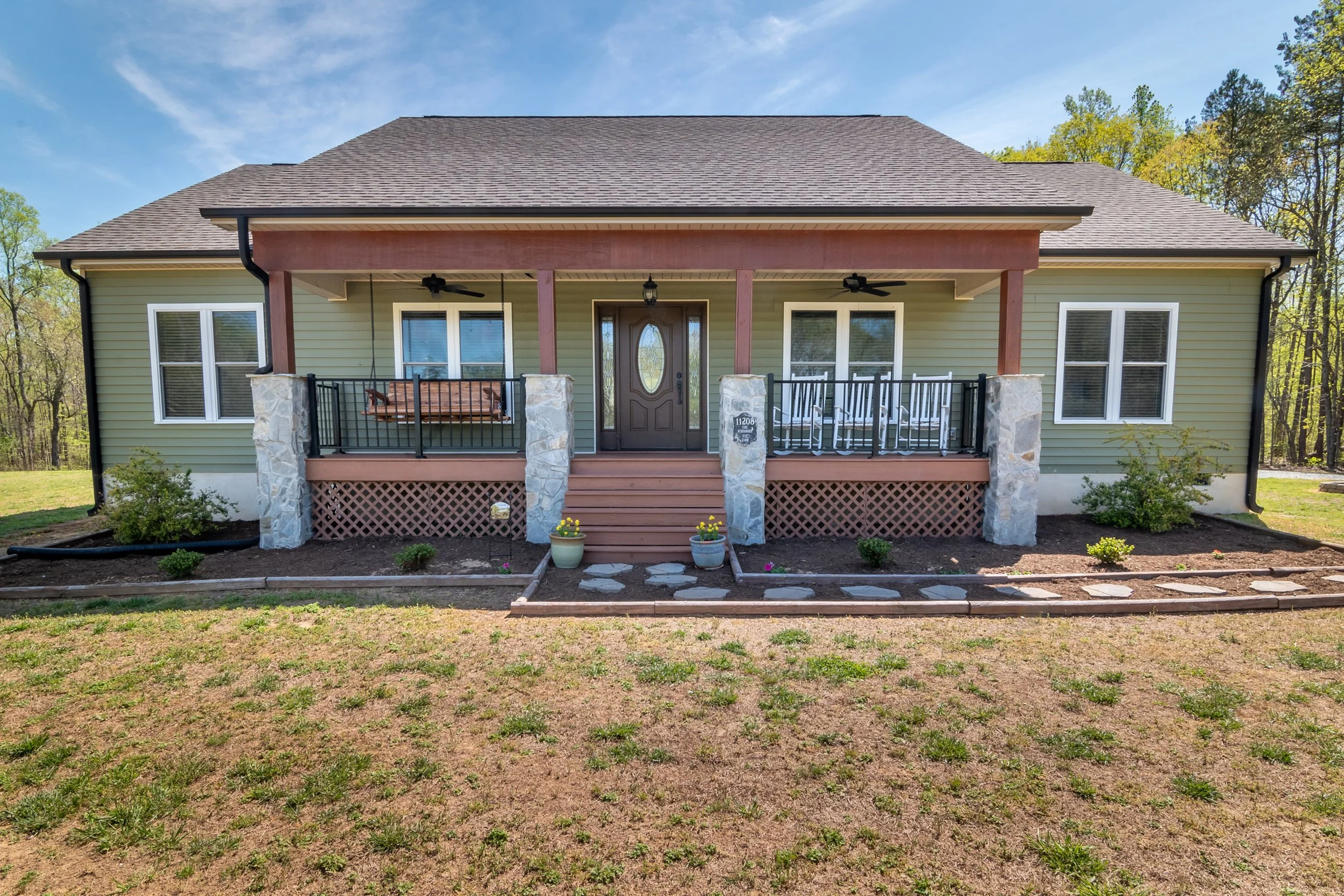 Front view of a single-story house with a green exterior, brown front door, and covered porch with stone pillars and railings. The porch has a swing and rocking chairs. Stone path leads to the entrance with plants and a lawn in the foreground.