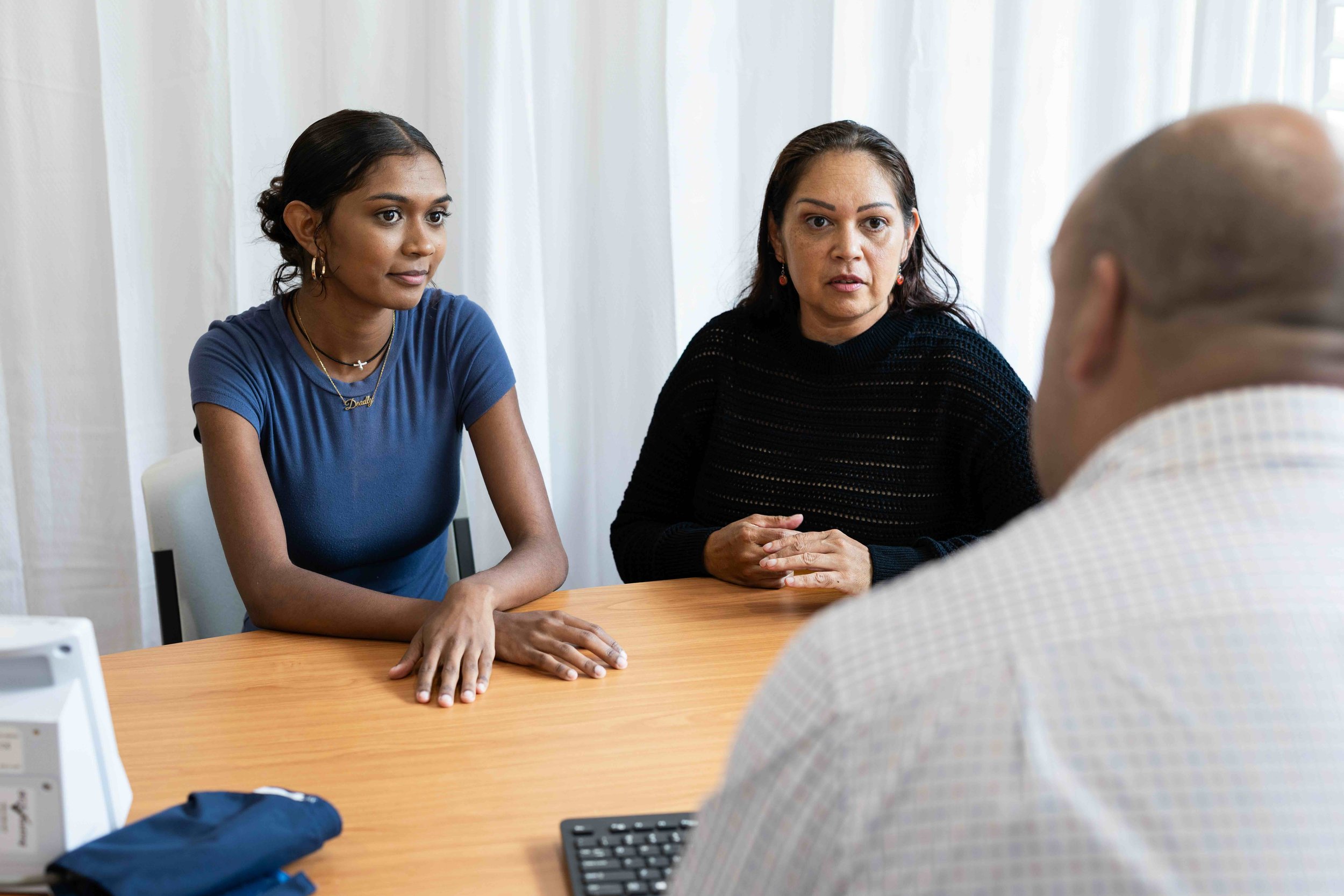 A young Aboriginal woman with dark brown skin and brown hair and an older Aboriginal woman with brown skin and brown hair sit at a desk talking to a man in a suit.