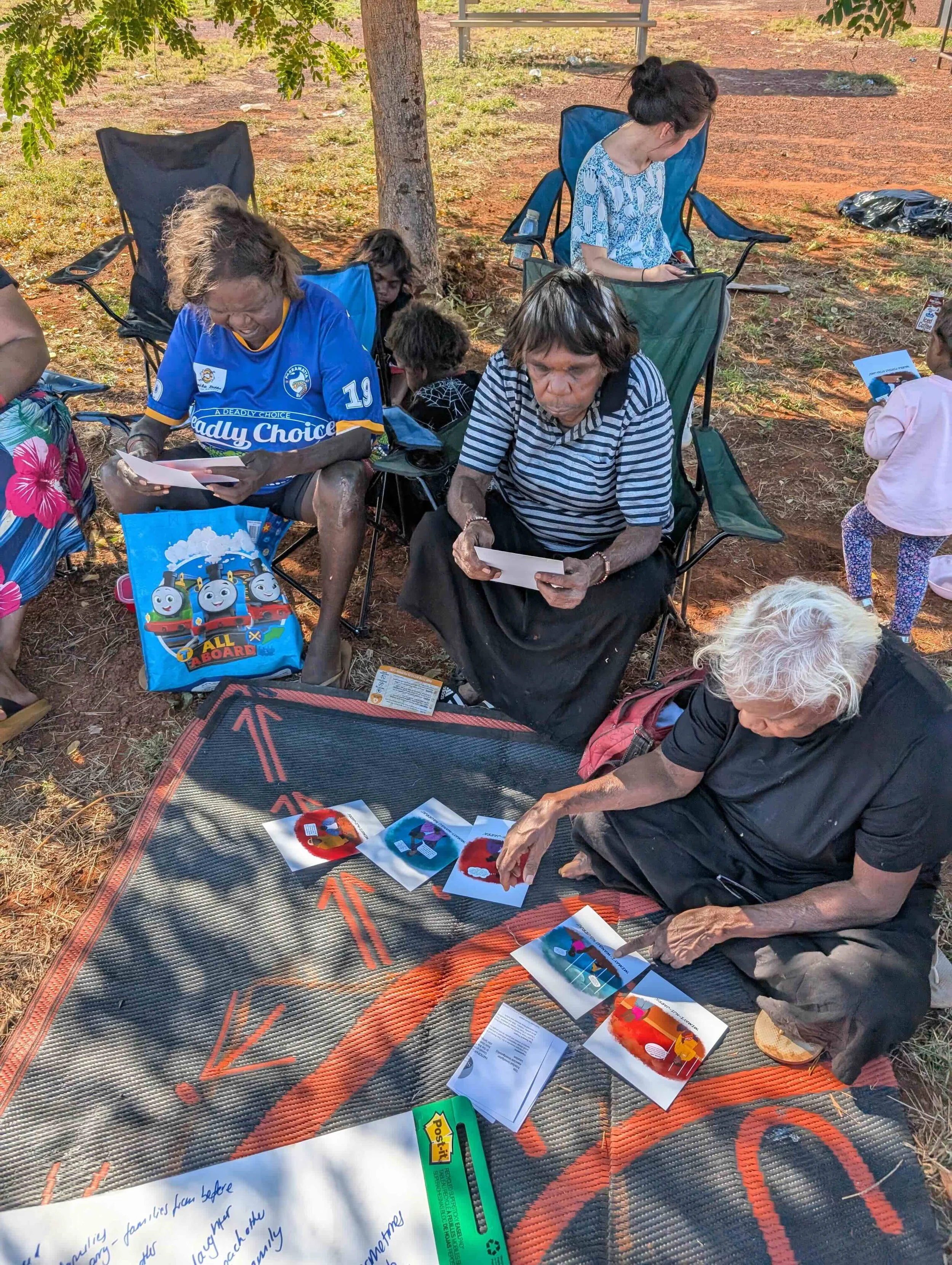 A group of older Aboriginal women sit on chairs and the ground looking at the Jealousing Cards, whilst children play around them. The cards have bright illustrations on them.