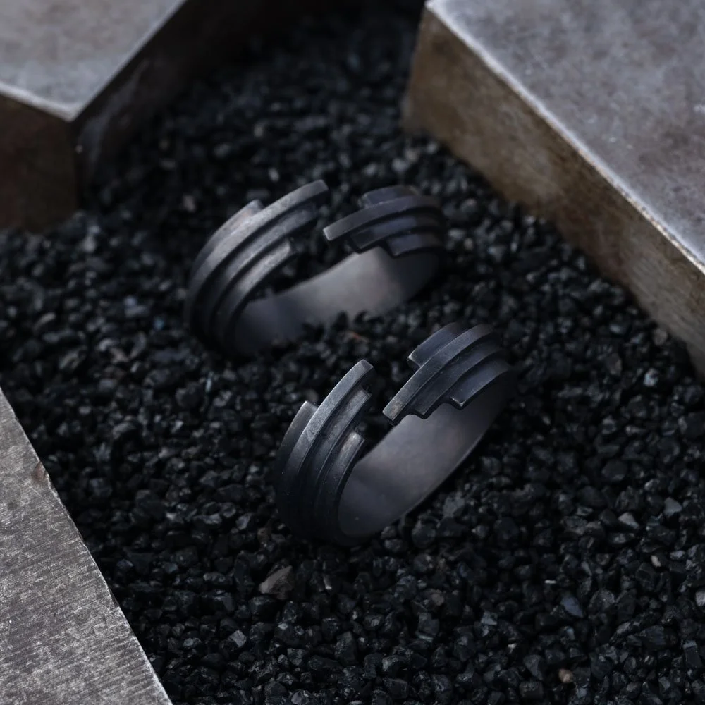 Two black rings from the 2073 City Decay Glitch collection, featuring stepped geometric cuts, placed on black coal