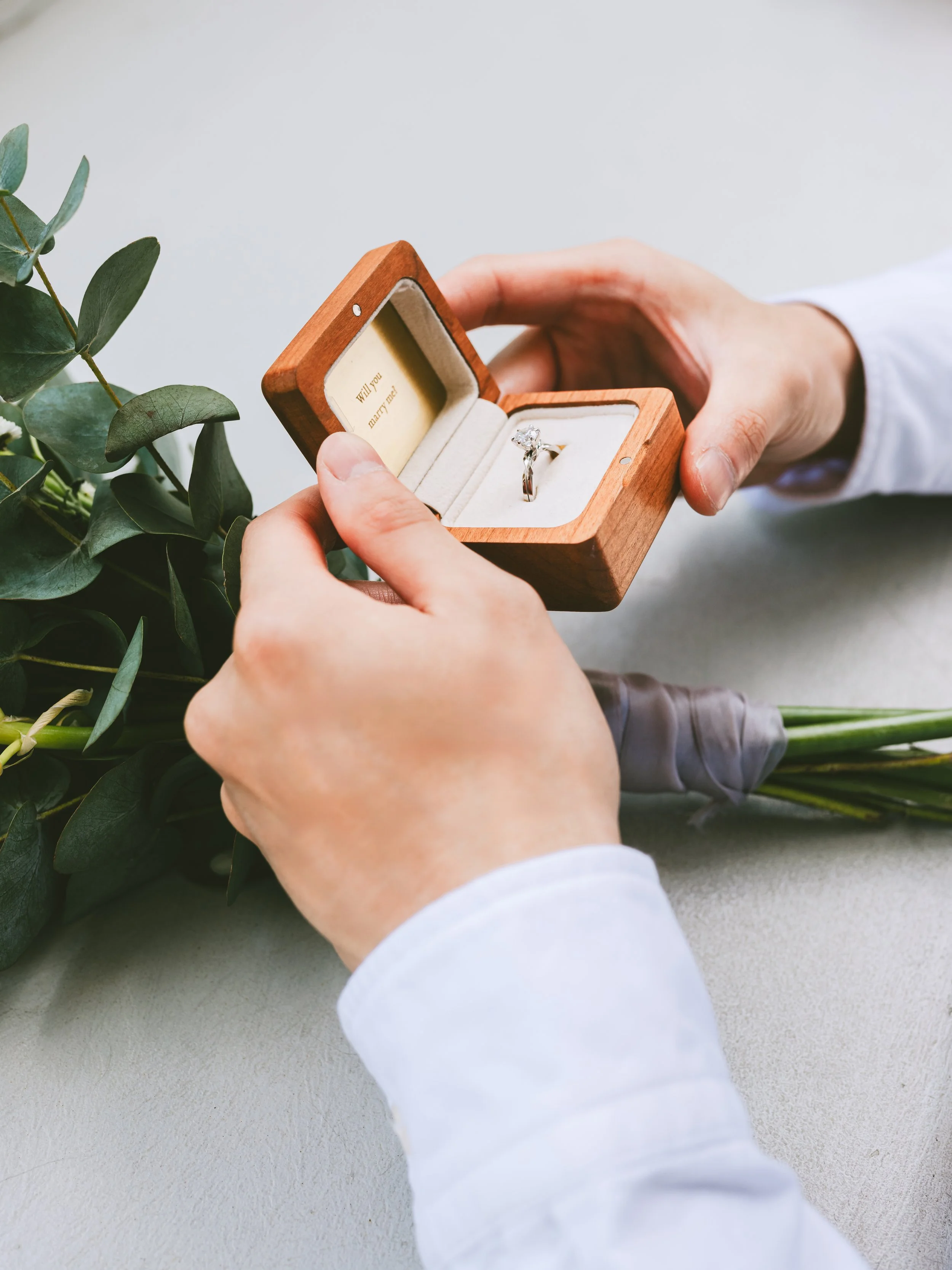 A man with white shirt holding a wooden ring box contain 18K Twin Souls Diamond Ring inside with a bouquet under the man's hand