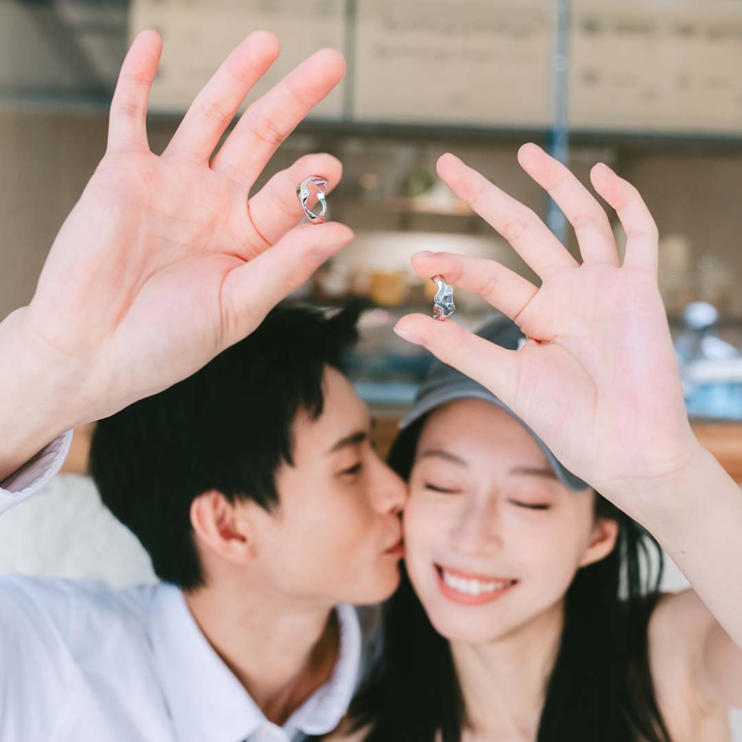 A couple holding born to meet collection wedding ring while a man in white shirt kissing the women on her cheek with long black hair wearing a cap