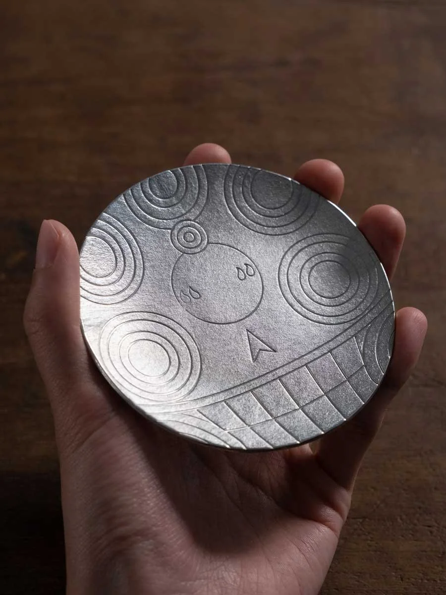 a hand holding a tin plate with Gudo face pattern on it