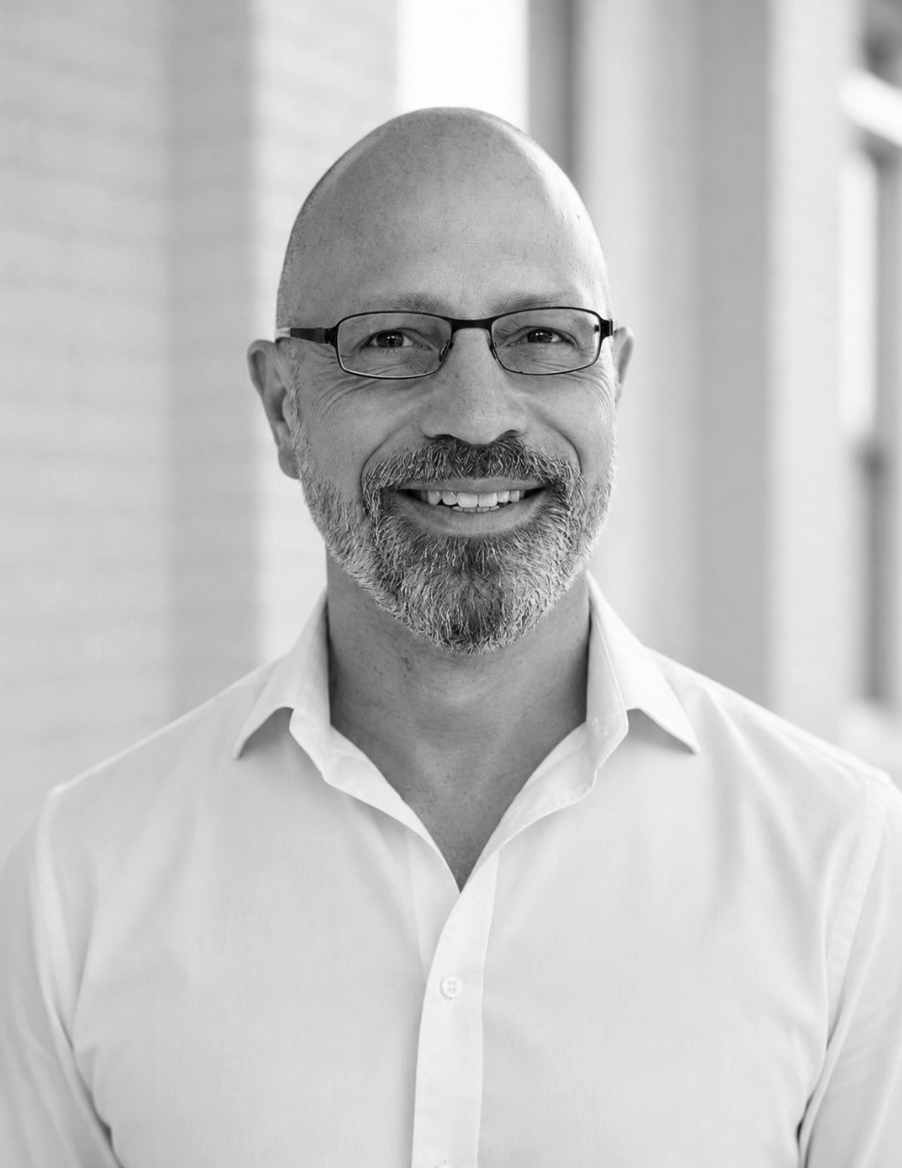 A smiling man with glasses, a beard, and a checkered shirt, against a plain background.