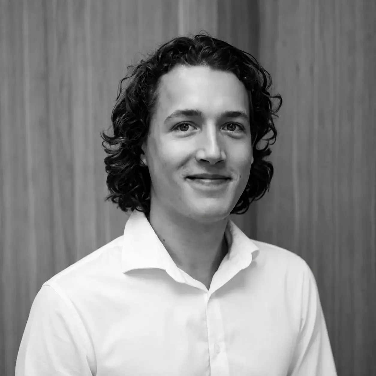 Black and white headshot of a young man with curly hair wearing a white collared shirt, smiling slightly, against a plain background.