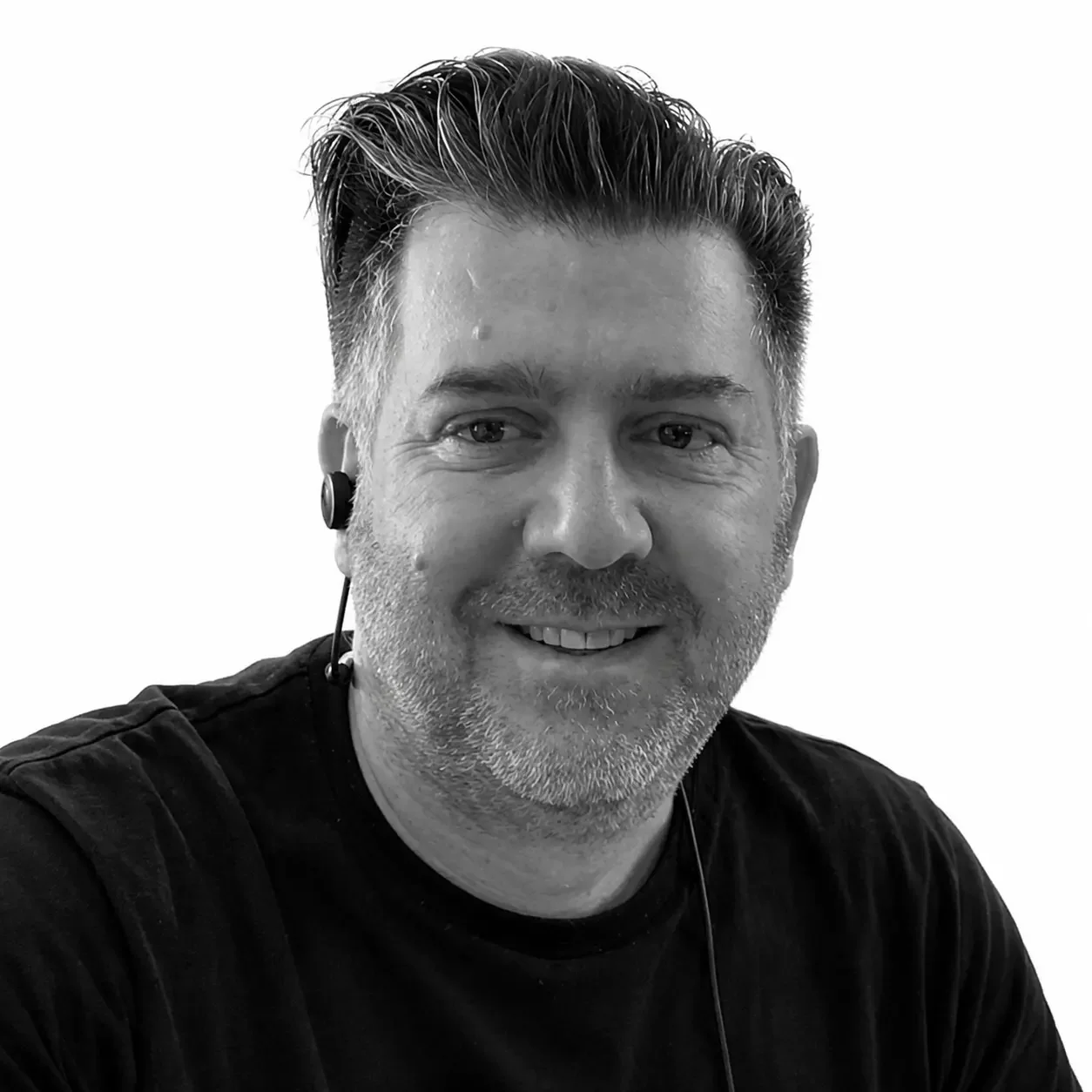 Black and white photo of a smiling man with short, styled hair, wearing earbuds, indoors near a window with visible trees outside.