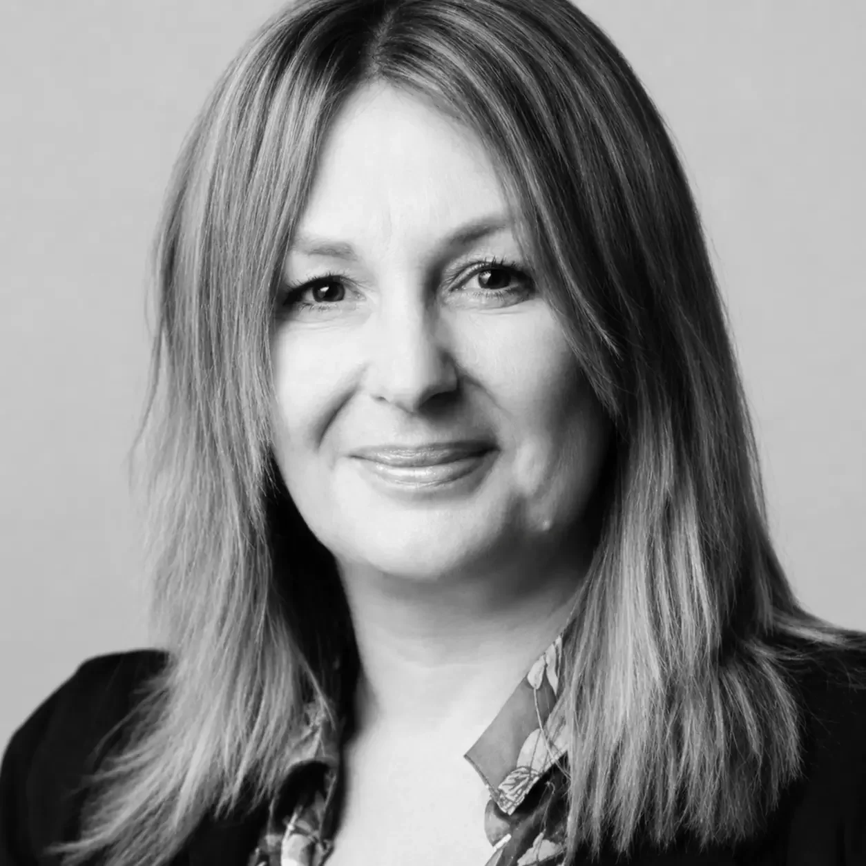 Black and white photo of a woman with long straight hair, smiling, wearing a dark blazer and patterned blouse.