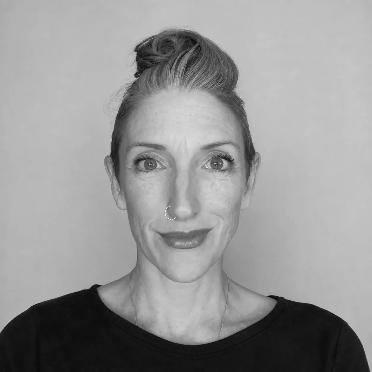 A woman with light skin and freckles, wearing a nose ring, has her hair tied up in a bun, and is smiling slightly. She is wearing a dark top and is photographed against a plain background.