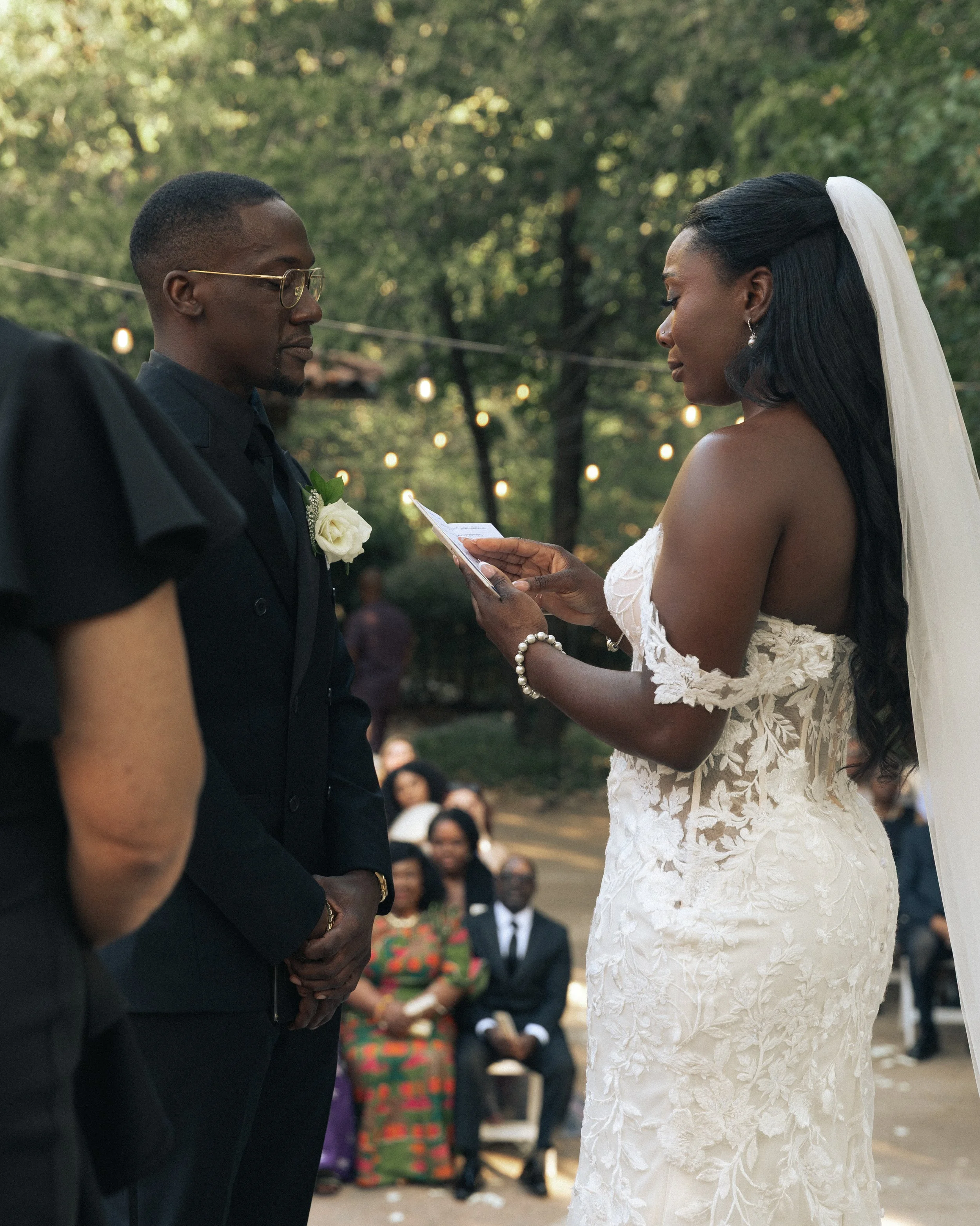 A bride and groom standing outdoors during a wedding ceremony, with the officiant reading vows. The bride is in a lace wedding dress with a veil, and the groom is in a black suit with glasses. Guests are seated in the background.