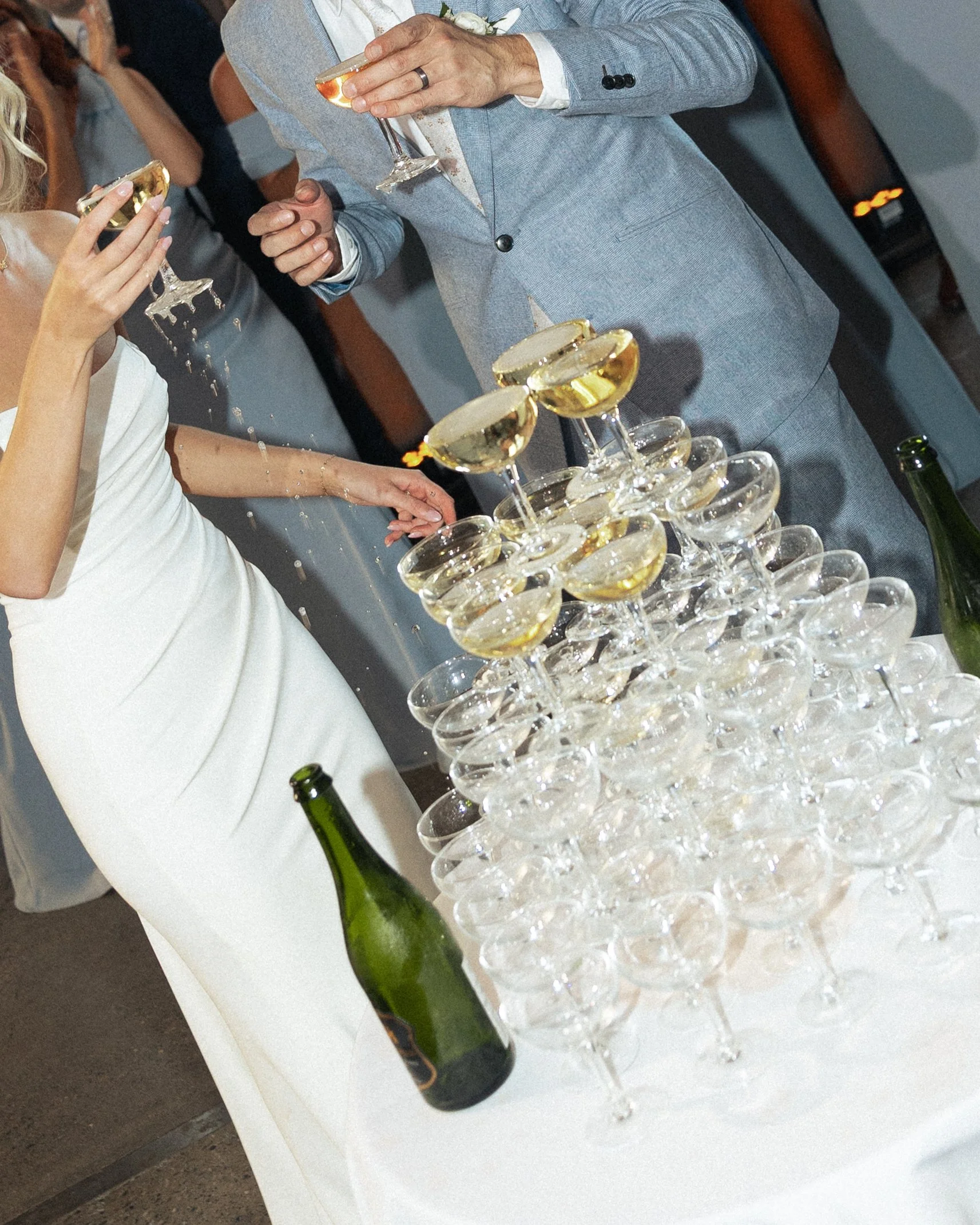 People celebrating with a Champagne tower at a wedding reception, with a woman pouring champagne and a man in a light gray suit.