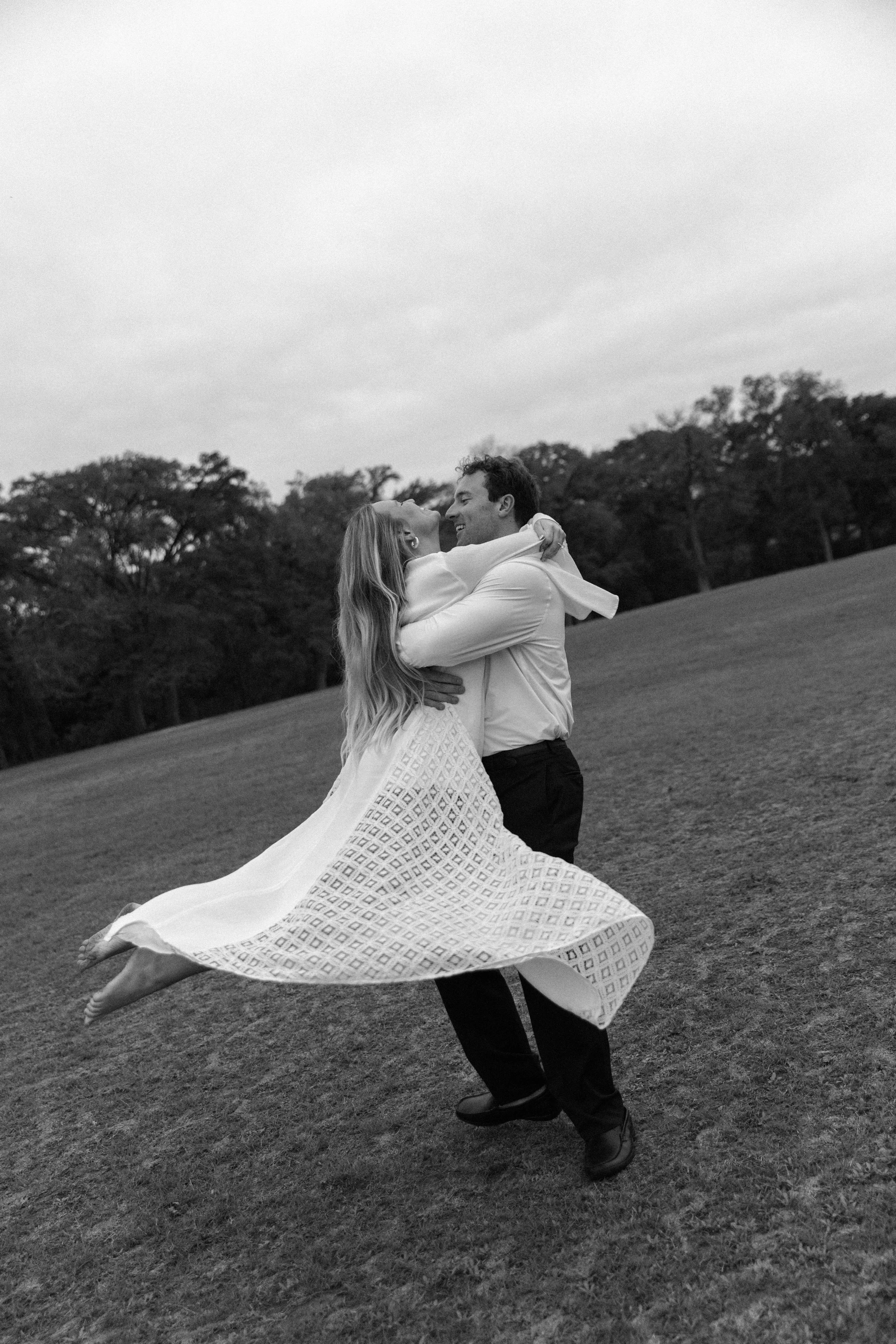 A couple dancing outdoors on a grassy hill, with the man lifting the woman as they embrace and smile, trees in the background, black and white photo.