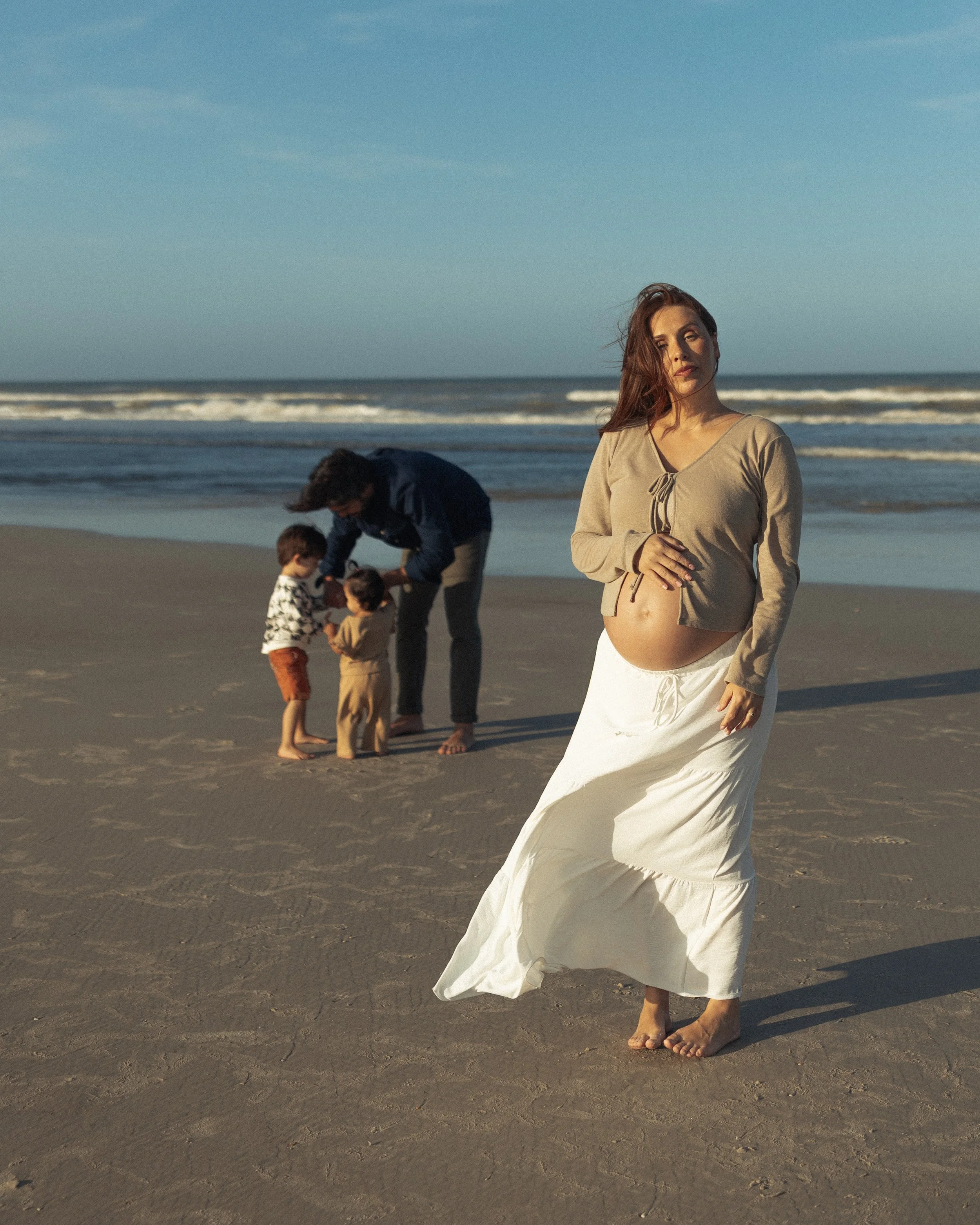 Pregnant woman standing barefoot on the beach with family in the background