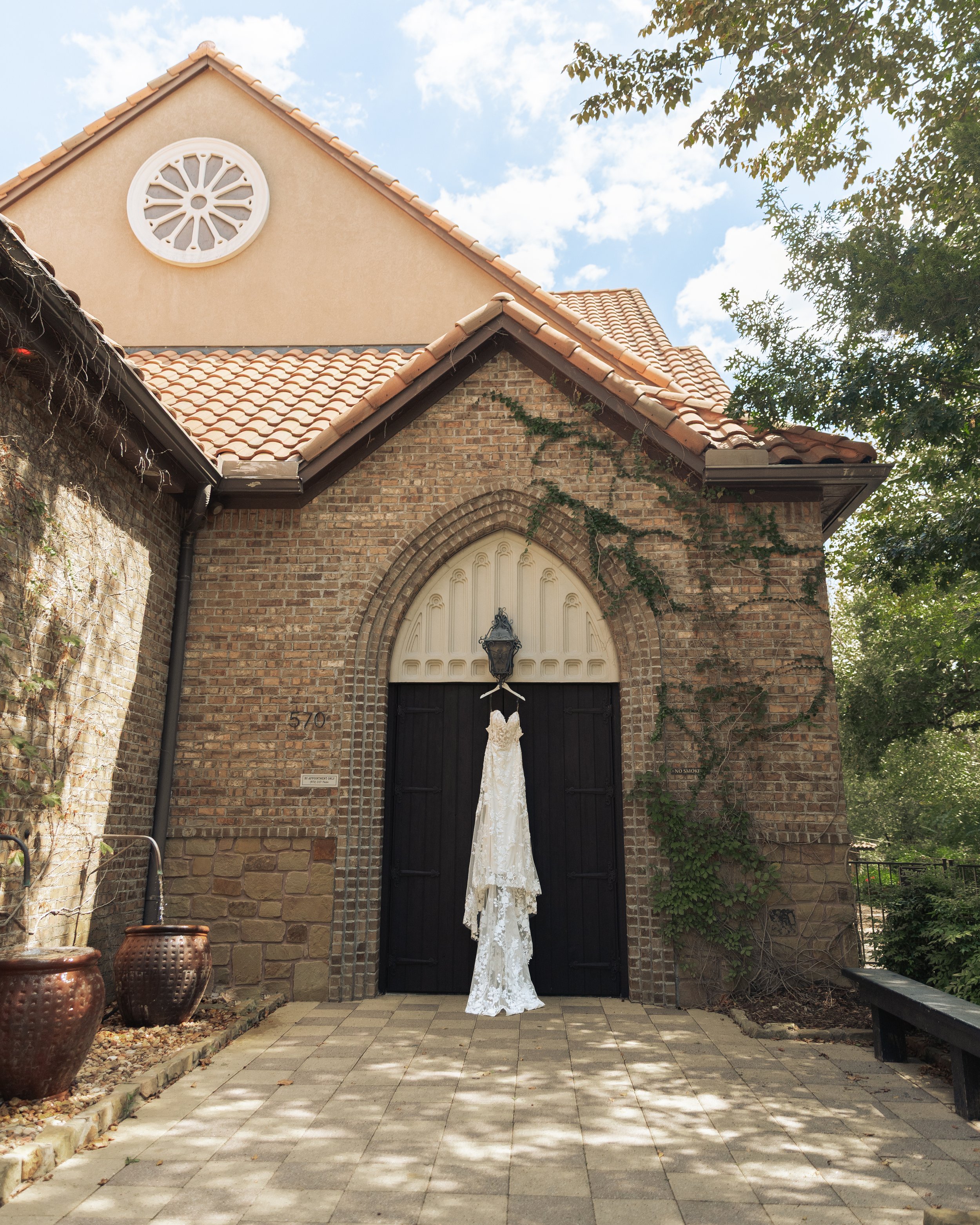 A wedding dress hanging on a doorway of a stone brick church with an arched entrance and a lantern above, surrounded by trees and a partly cloudy sky.