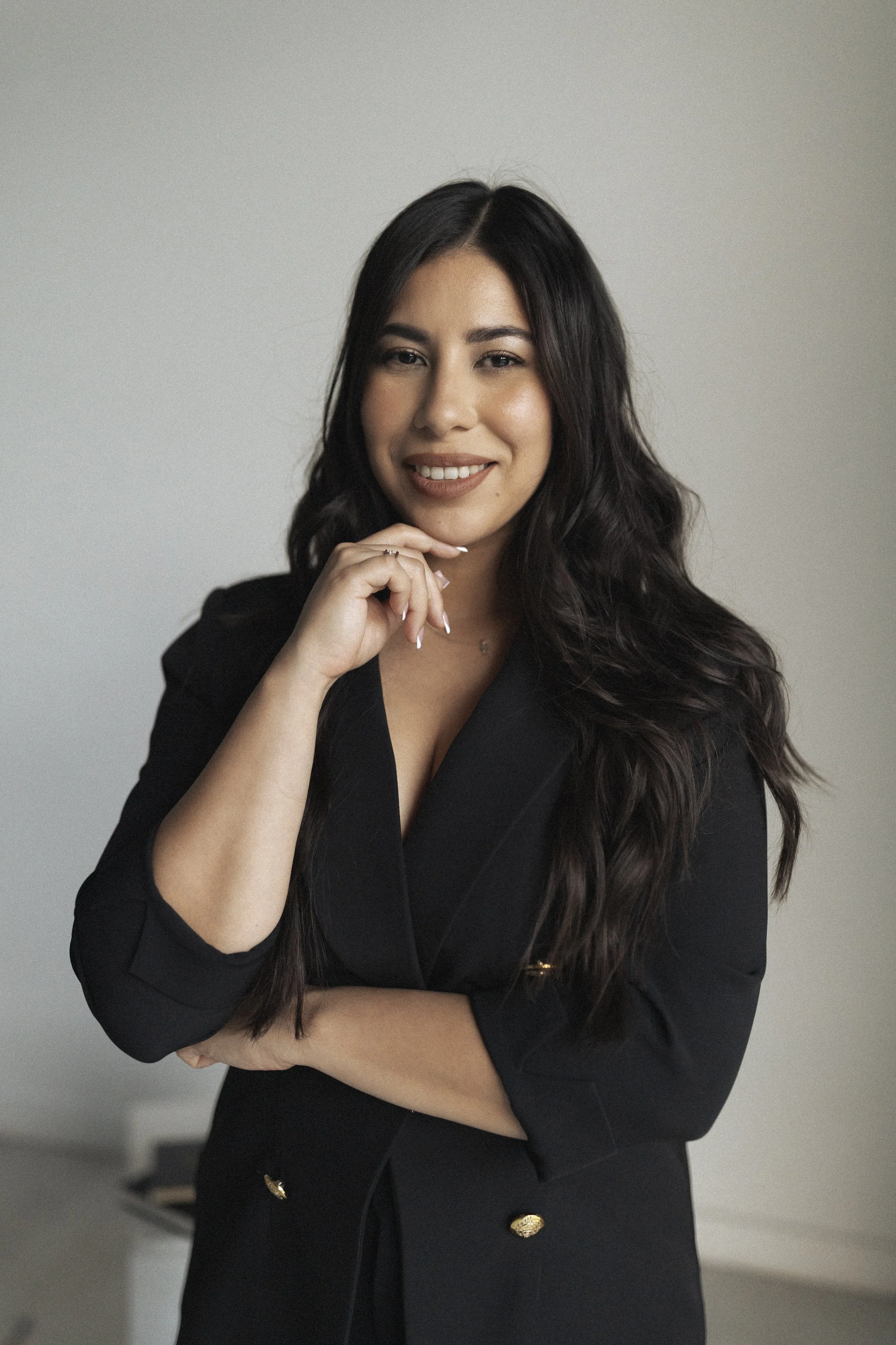 A young woman with long, dark wavy hair, wearing a black blazer, smiling and resting her chin on her hand, standing in front of a plain light-colored wall.