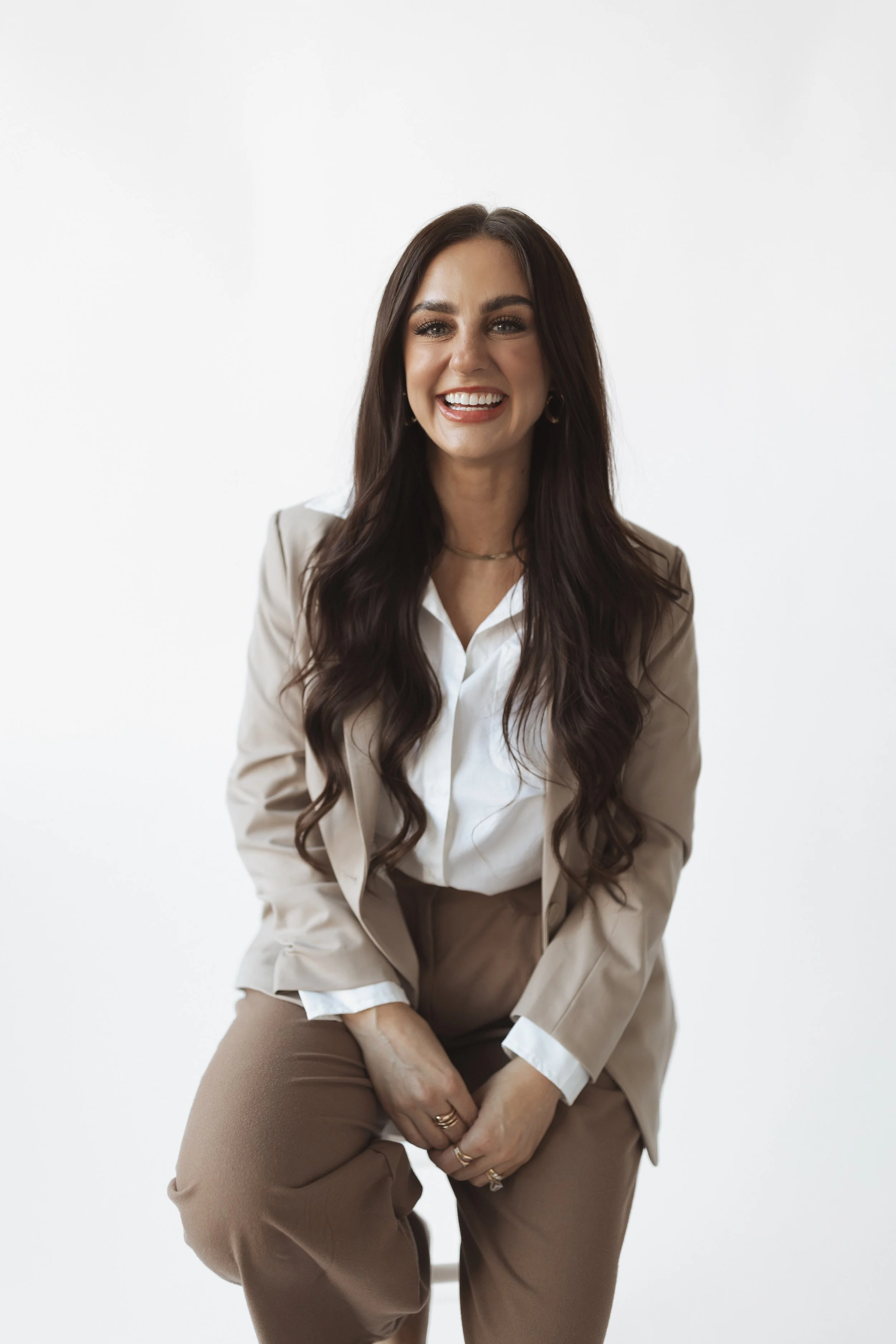 A woman with long dark hair, smiling and wearing a beige blazer, white shirt, and beige pants, sitting against a plain white background.