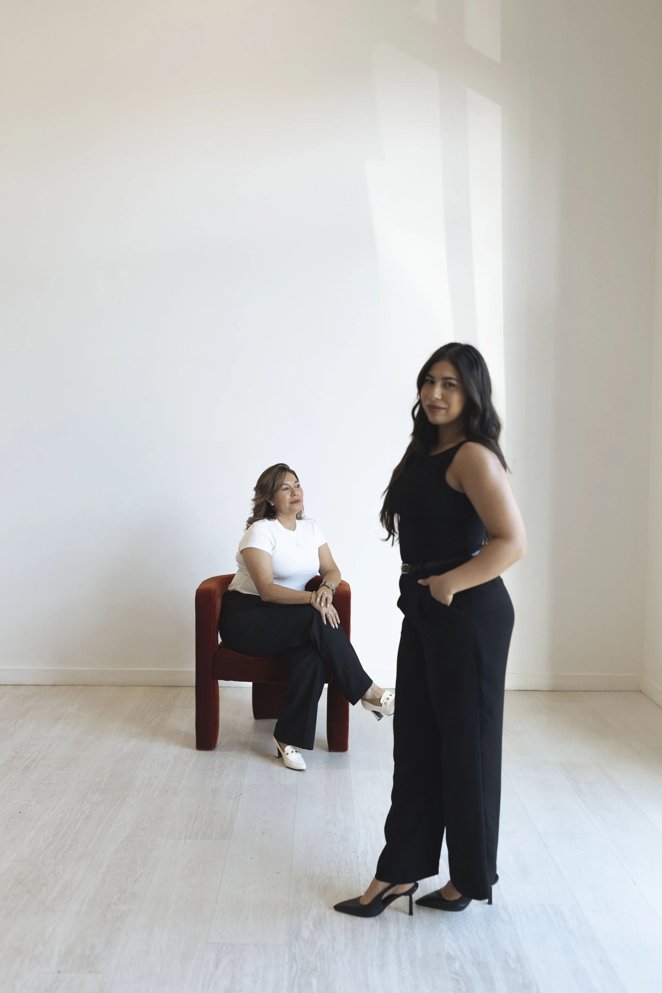 Two women in black and white outfits, one sitting on a red chair and the other standing, in a minimalist room with white walls and light-colored wooden flooring.