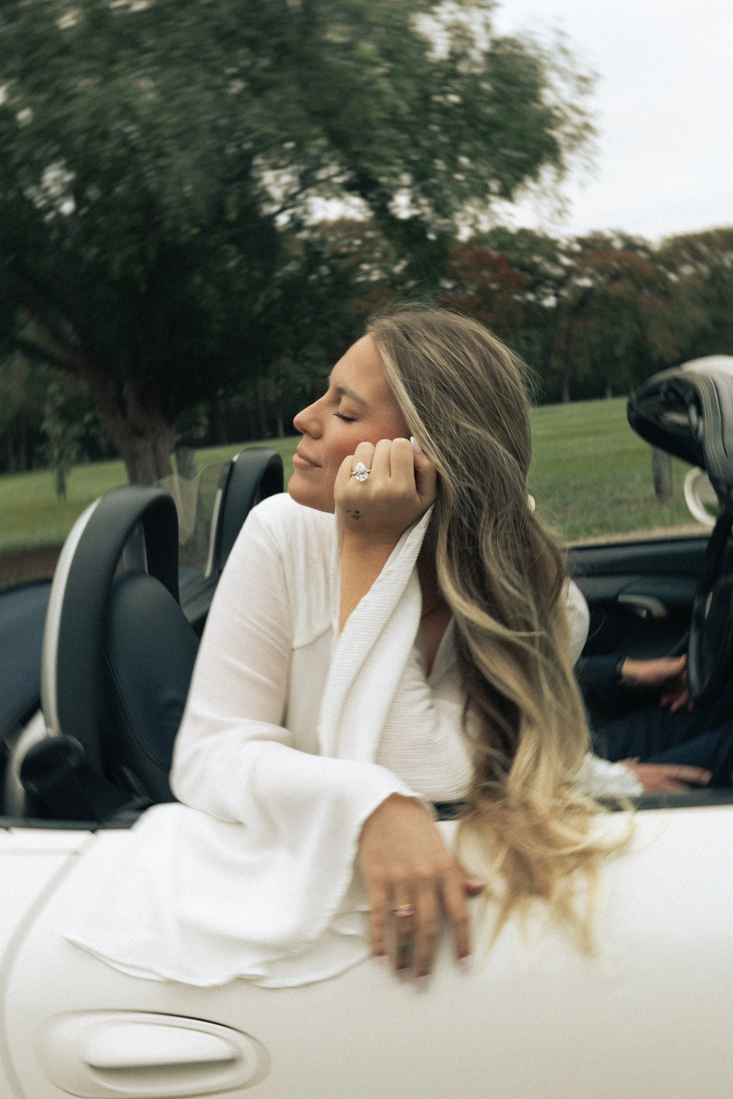 A woman with long wavy hair, wearing a white outfit, and a large ring, sits in a convertible car with her eyes closed, relaxing with her head tilted to the side, in a park with trees in the background.