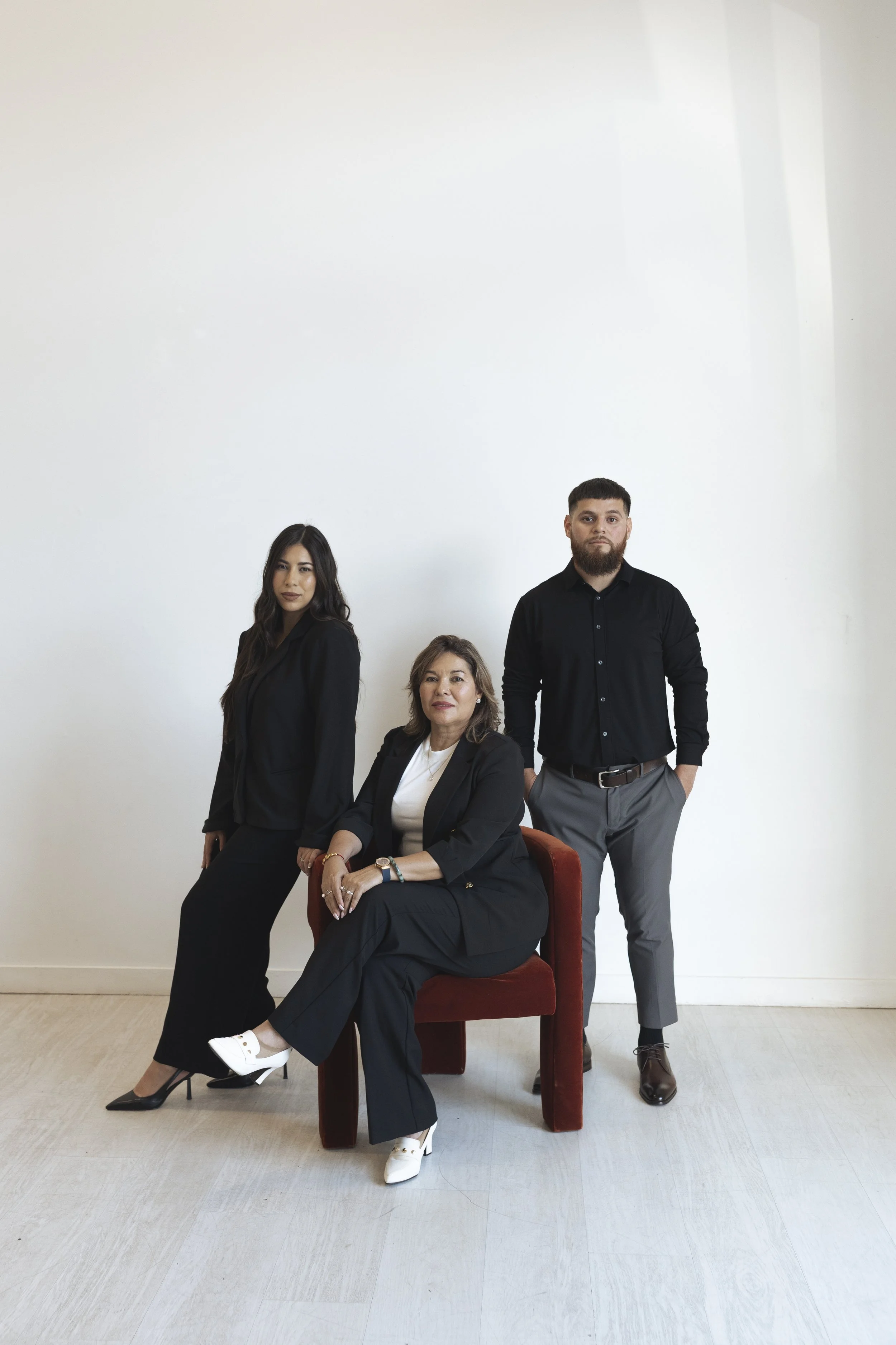 Three professionally dressed people posing against a plain white wall, seated on and standing around a red chair.