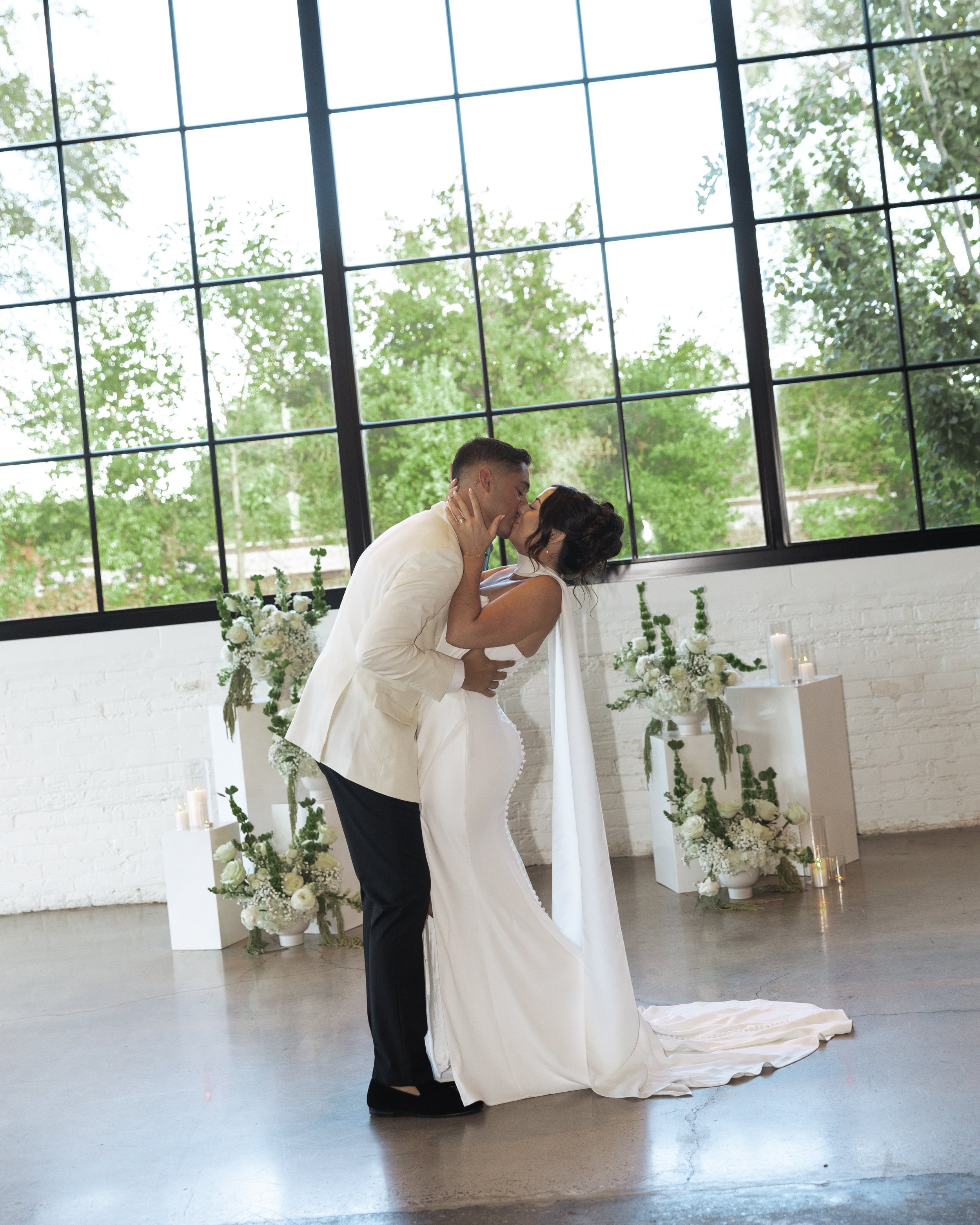 A couple shares a kiss during their wedding ceremony in front of large windows with greenery outside, decorated with white flowers and candles.