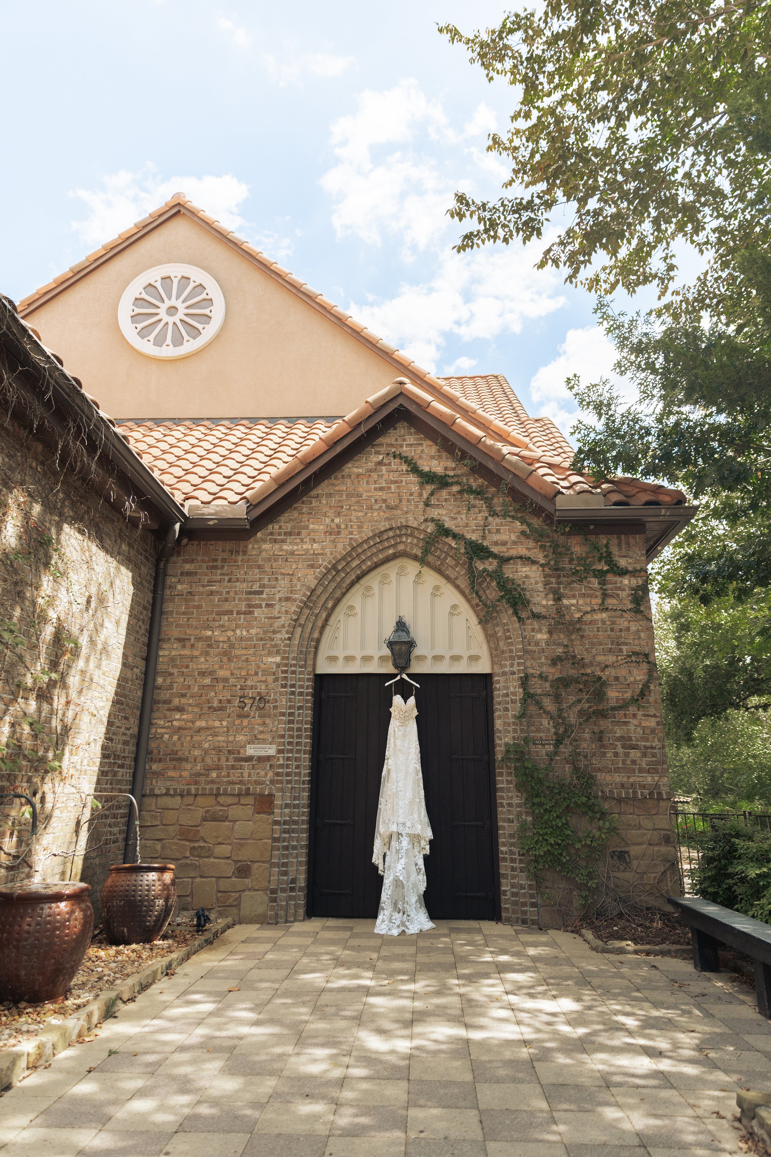 A white lace wedding dress hangs on a hanger from a lamp post in front of a dark wooden church door. The church has brick and stucco walls, a tiled roof, ivy growing on the wall, and a round window on the upper part of the building. The ground is pav