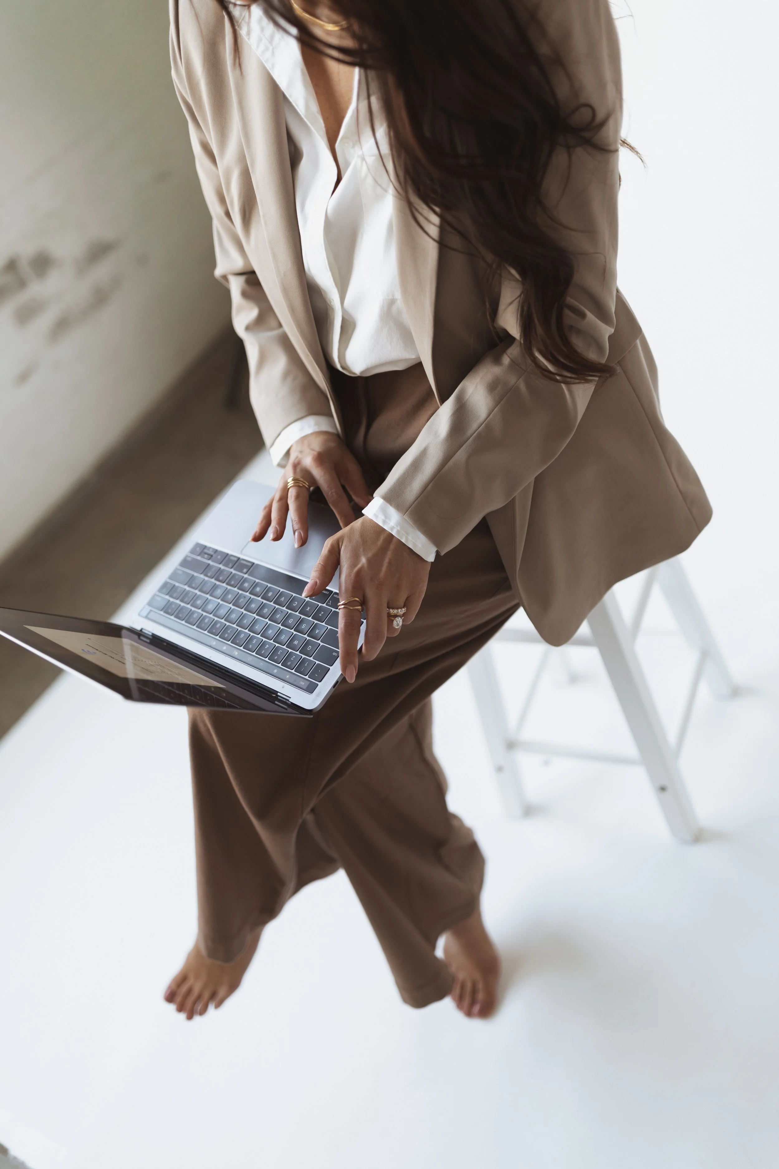 A woman in beige suit and white shirt using a laptop while standing barefoot on a white surface.