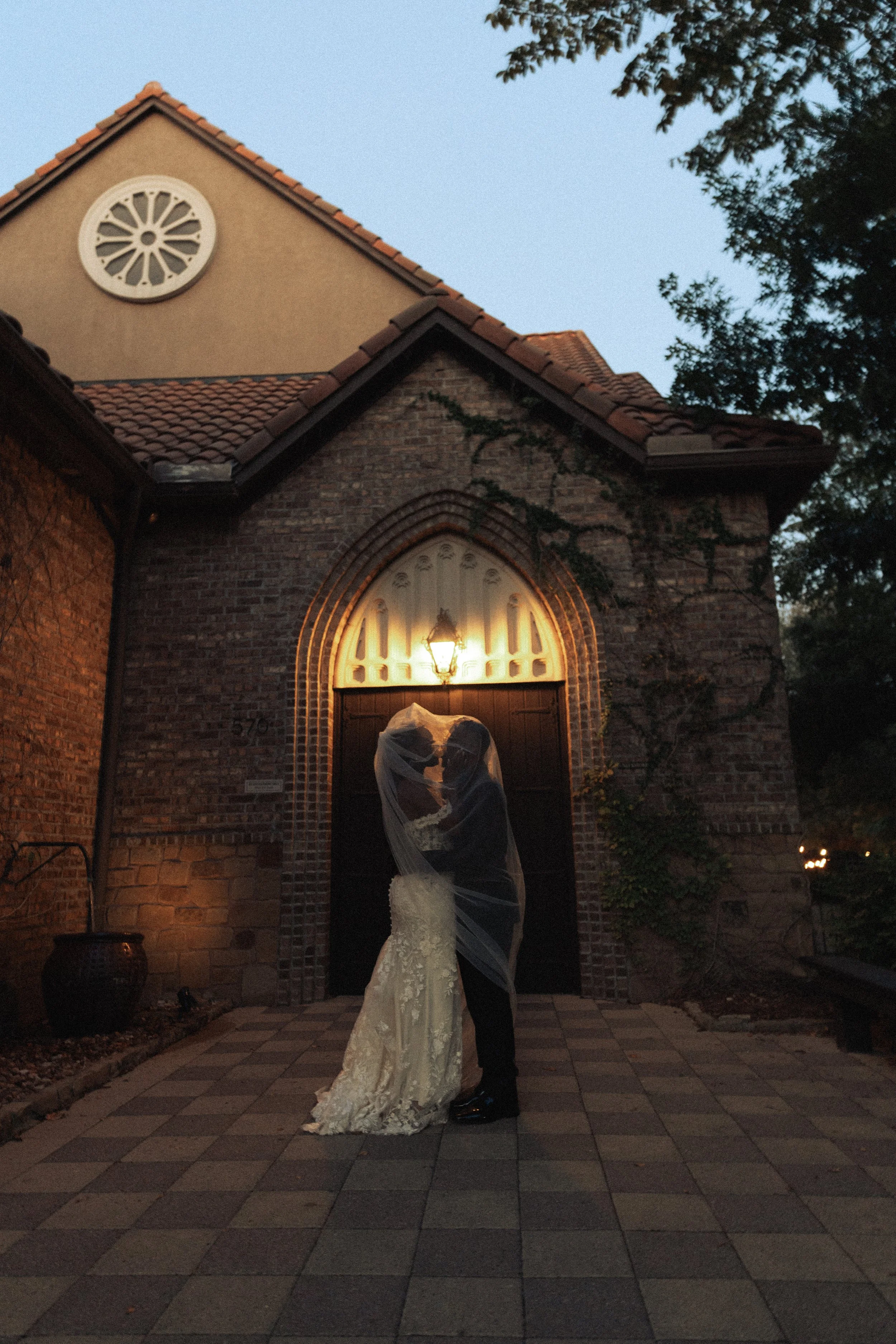 A bride and groom kissing under a veil outside a brick church at dusk.