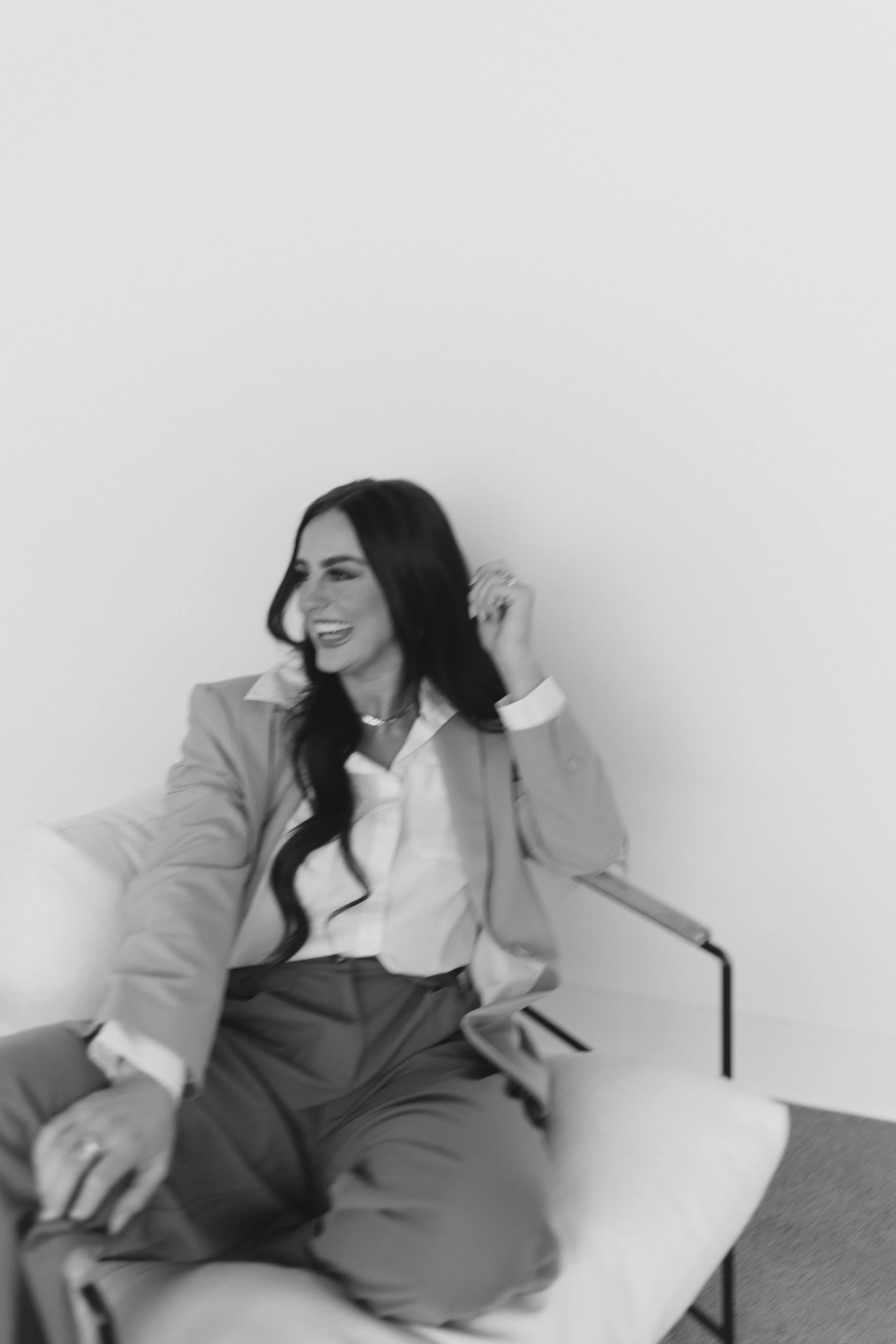 A woman sitting on a light-colored sofa, smiling and looking to the side, dressed in a light blazer, white shirt, and dark pants, with long dark hair.