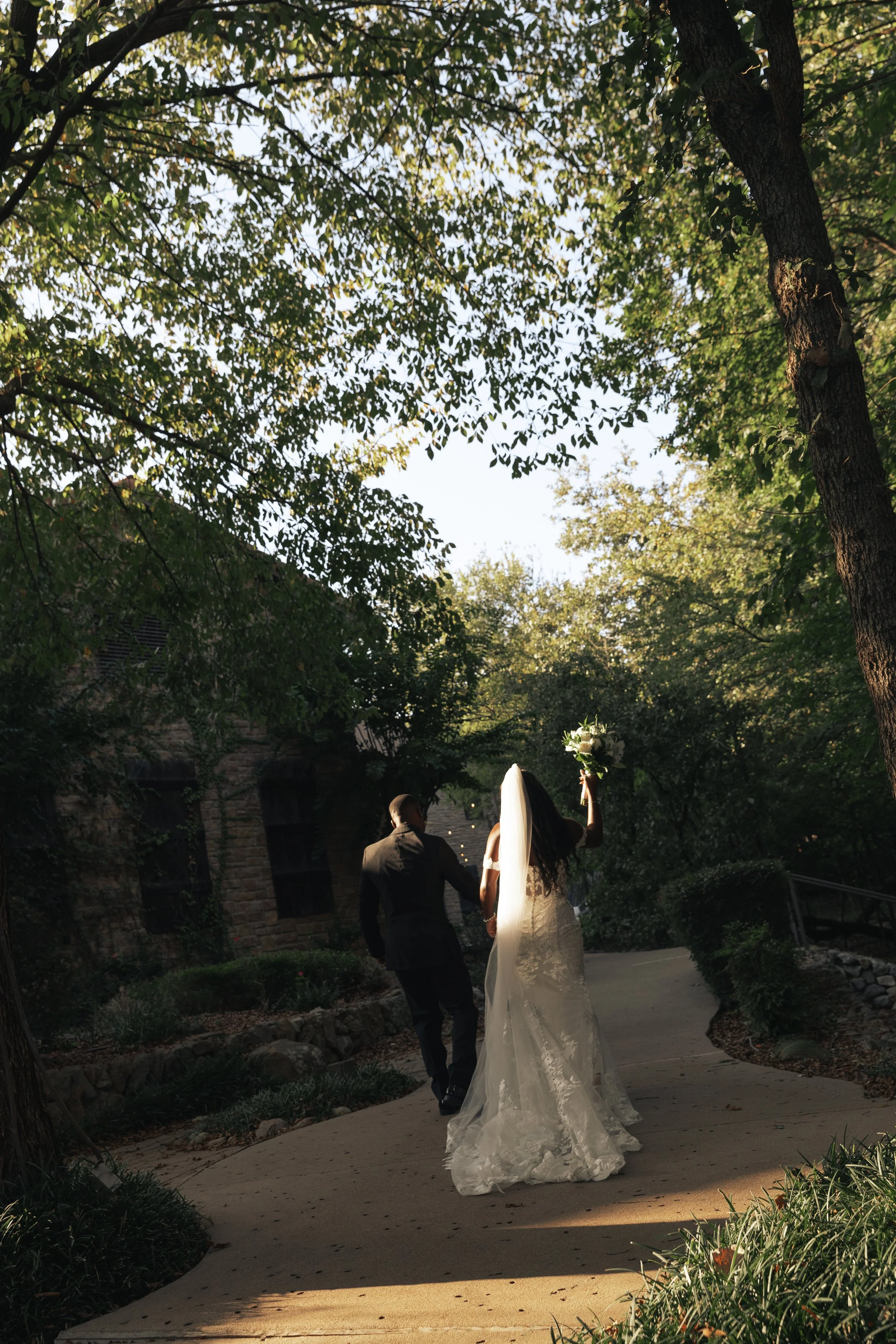 A bride in a white wedding dress and veil walking with a groom in a suit along a winding outdoor path surrounded by trees, with the bride holding a bouquet and raising her arm.