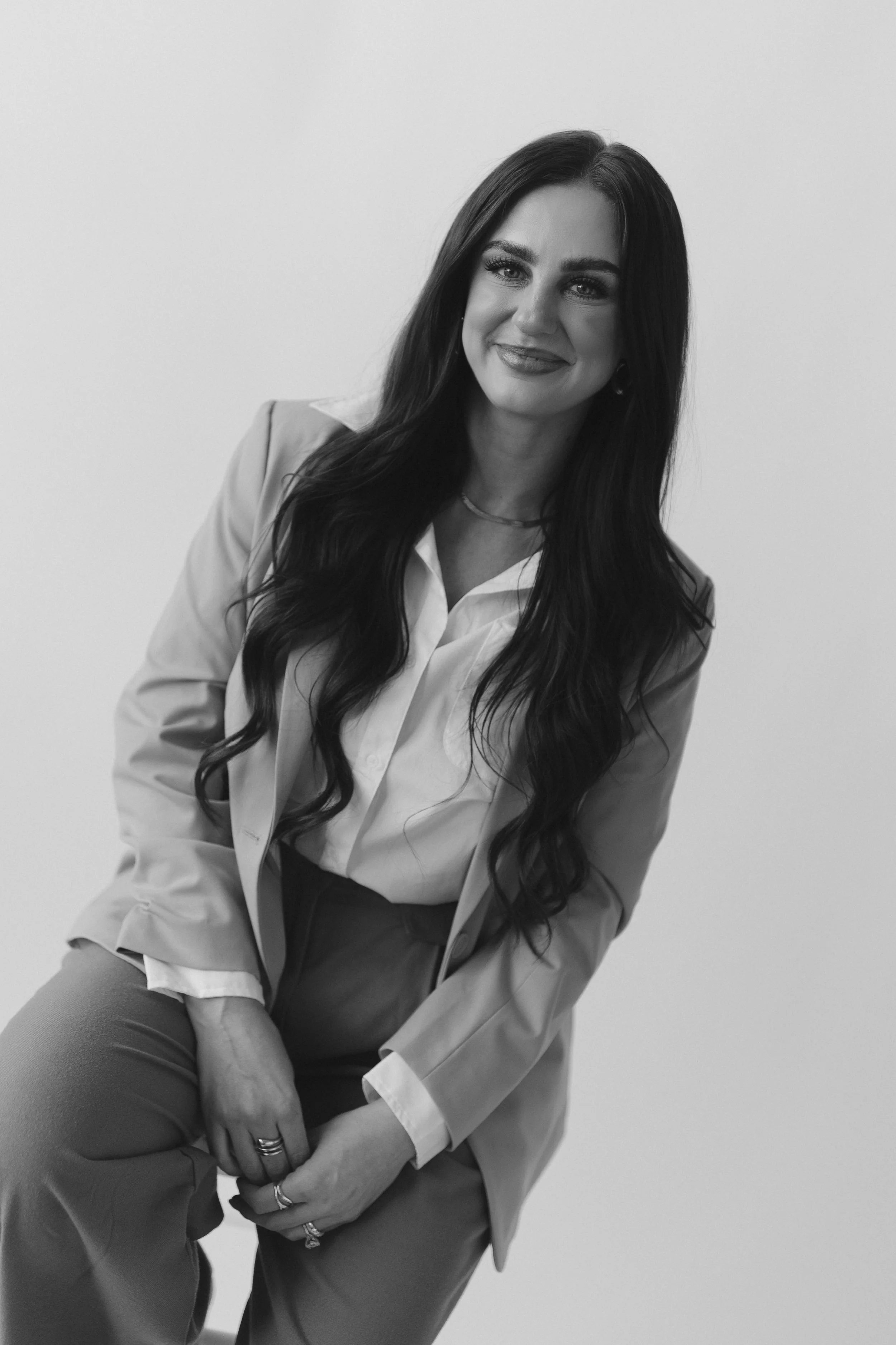Black and white photo of a woman with long wavy hair wearing a blazer and trousers, smiling and sitting against a plain background.