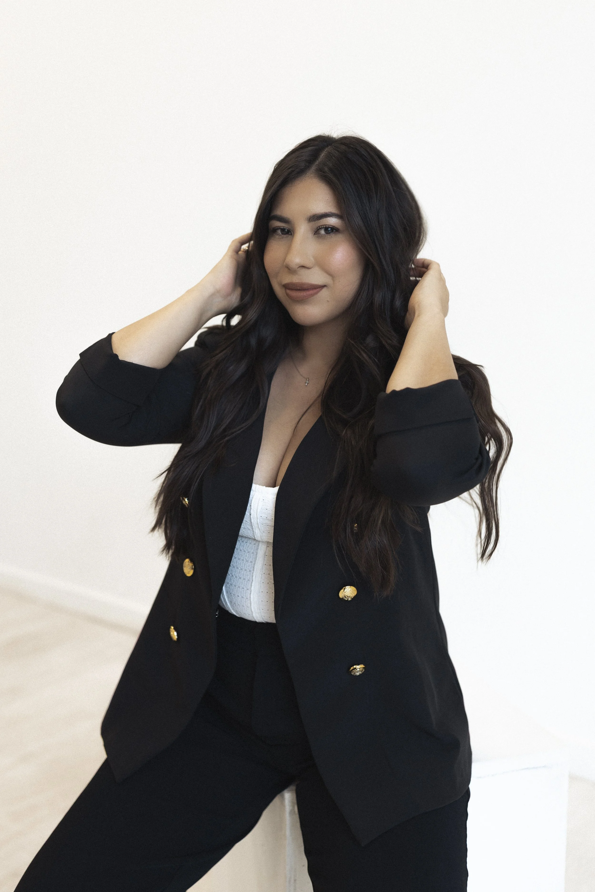 A woman with long dark hair wearing a black blazer with gold buttons, a white top, and black pants, posing indoors against a plain white wall.