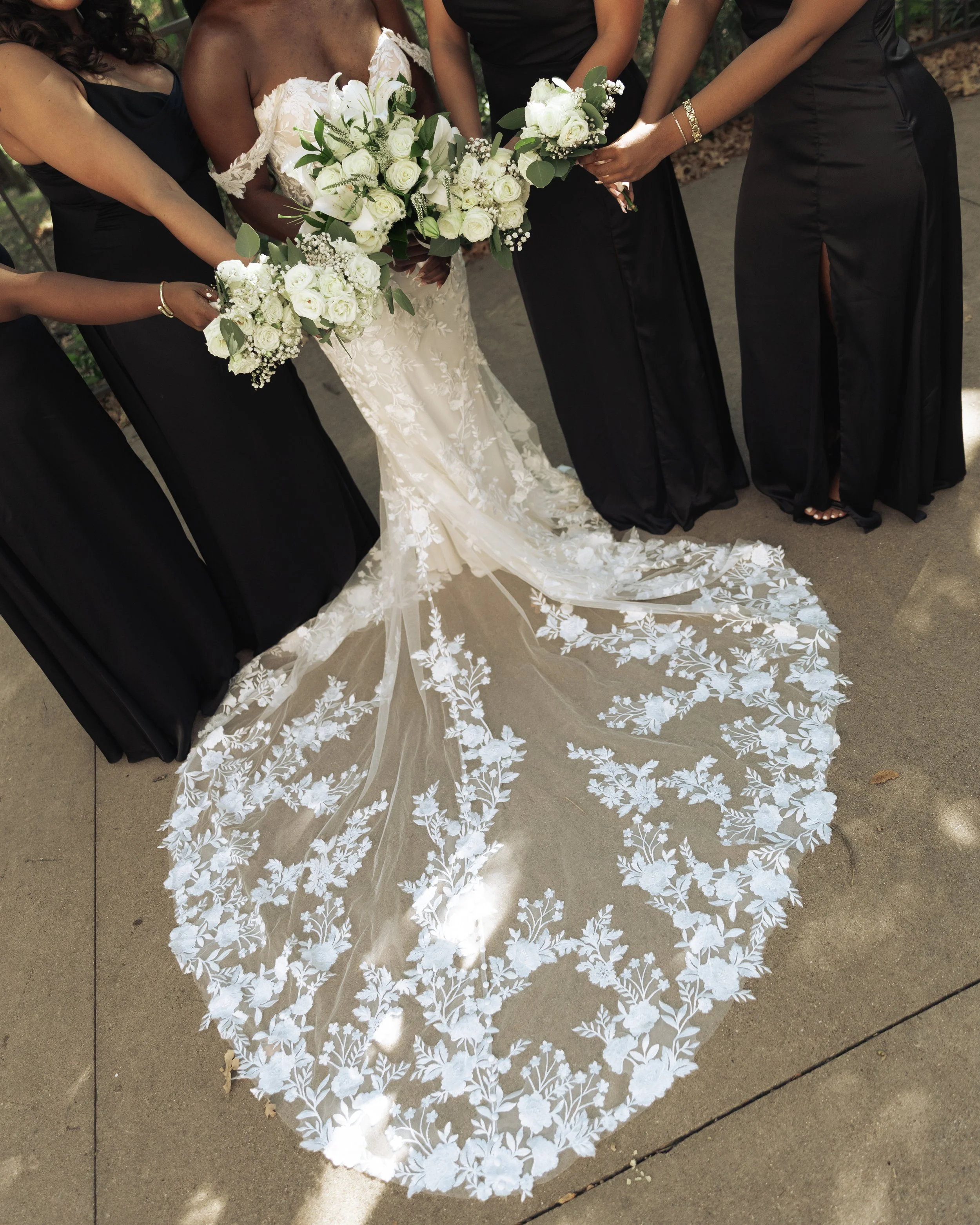 Bride in a white wedding gown and veil holding a bouquet of white roses, surrounded by four women in black dresses holding bouquets, outdoors on a concrete surface.