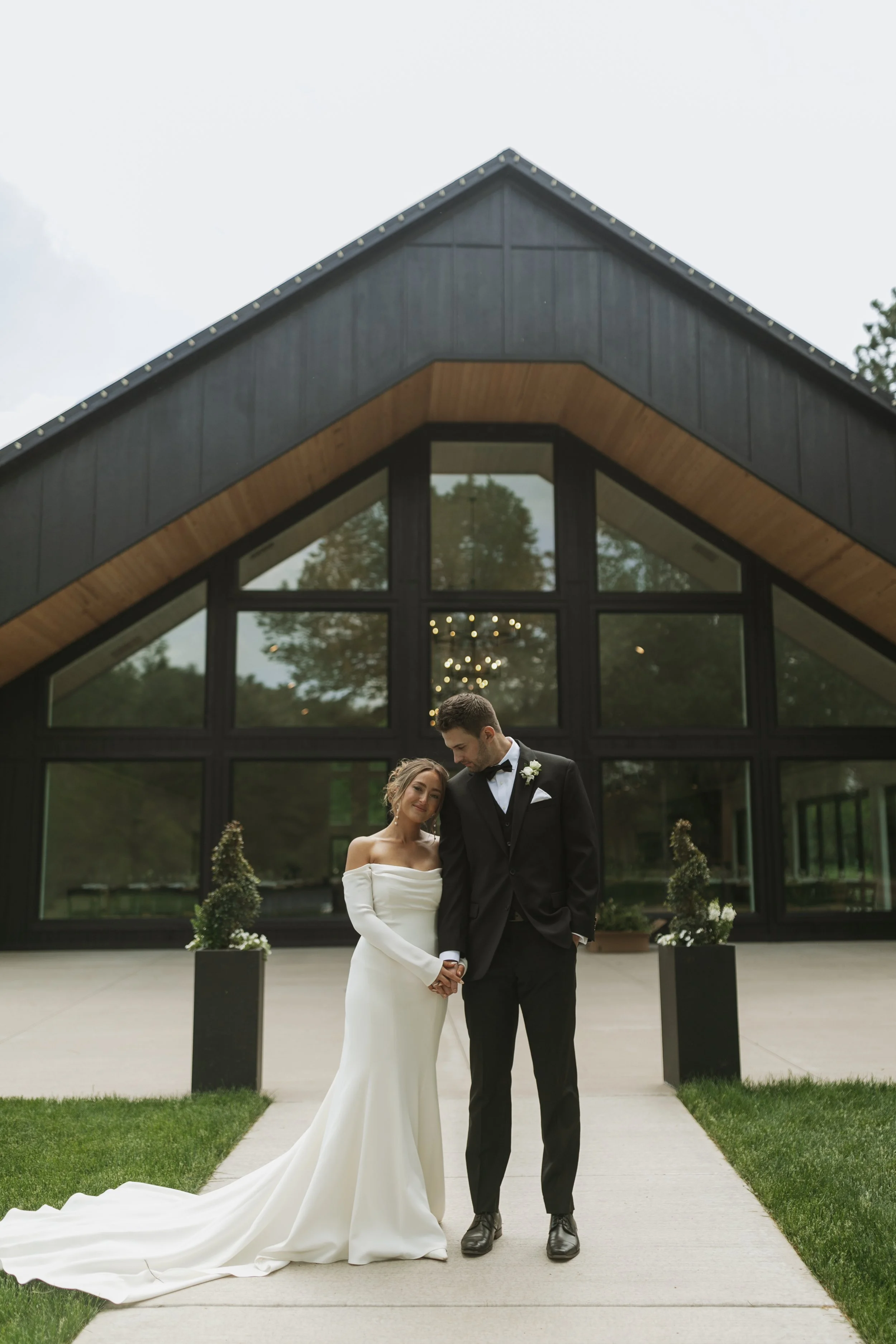 Bride and groom holding hands outside a modern church during their wedding, with the bride in a white off-shoulder gown and the groom in a black tuxedo, in front of large glass windows.