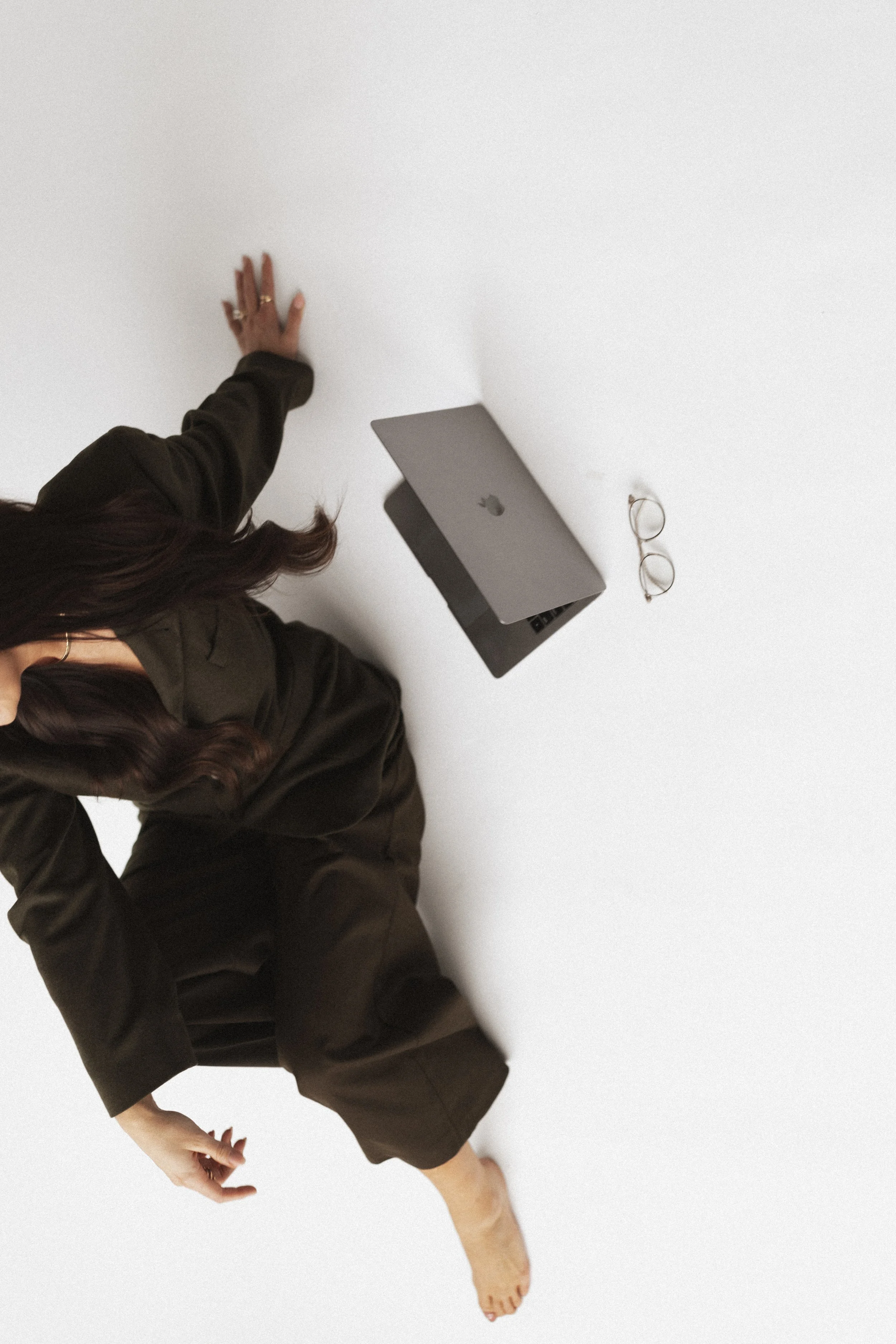 A person in dark clothing with long hair, sitting barefoot on the floor, with a laptop, glasses, and a hand behind their head.