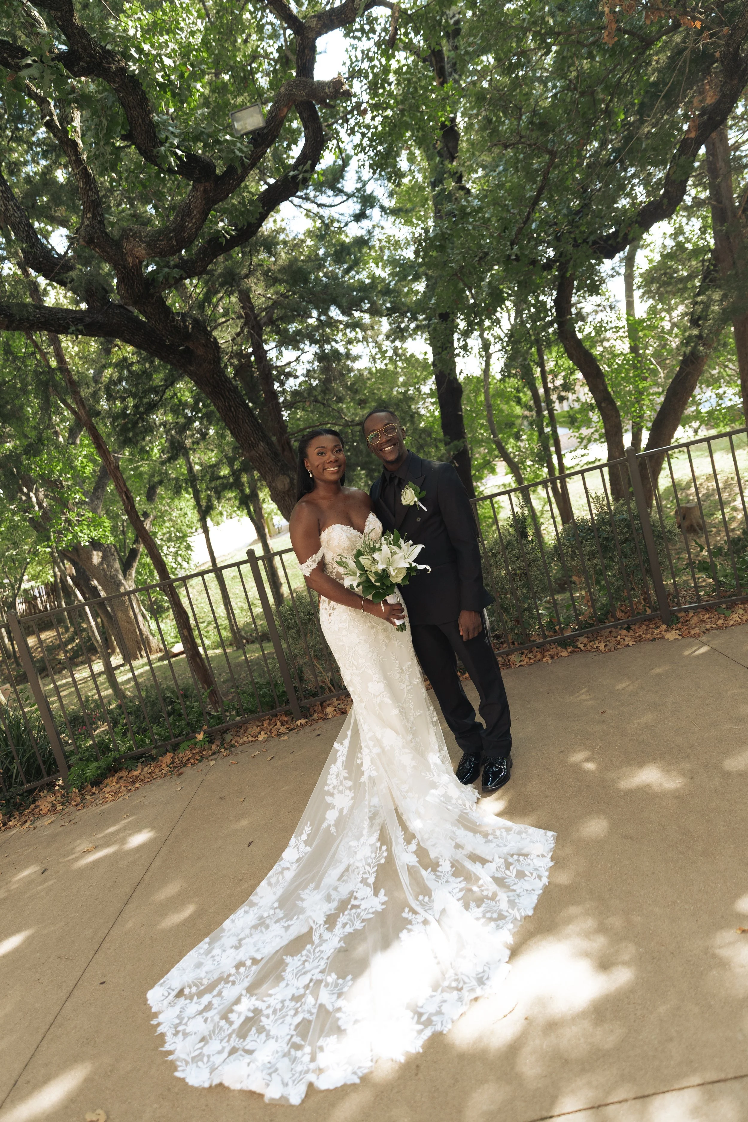 A newlywed couple standing outside under a large leafy tree with a metal fence in the background. The bride wears a white lace wedding gown and holds a bouquet of white lilies and greenery. The groom wears a black suit with a white boutonniere. Both 
