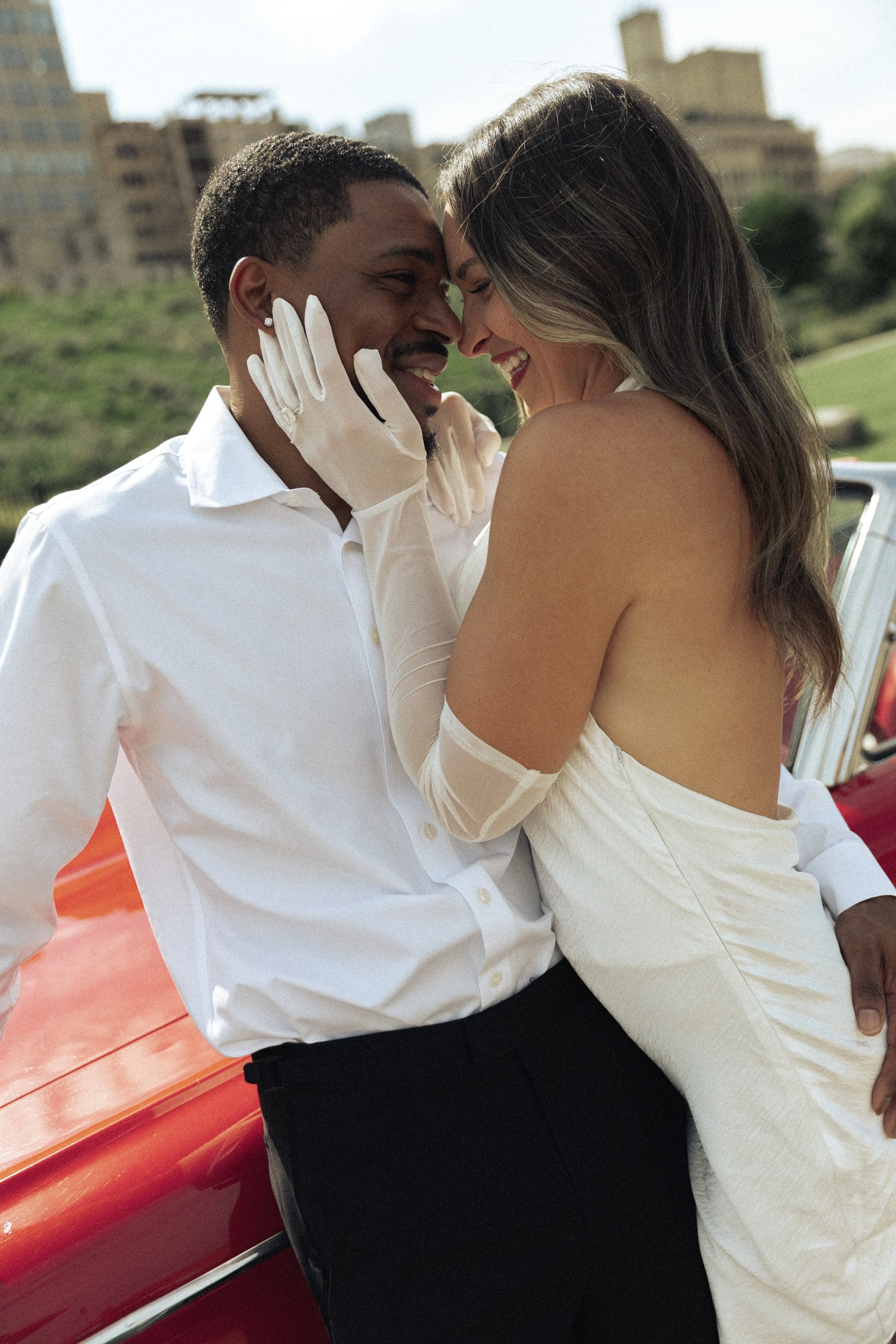 A happy couple with the woman in a white dress and the man in a white shirt, bonding closely outdoors near a red vintage car with a cityscape in the background.