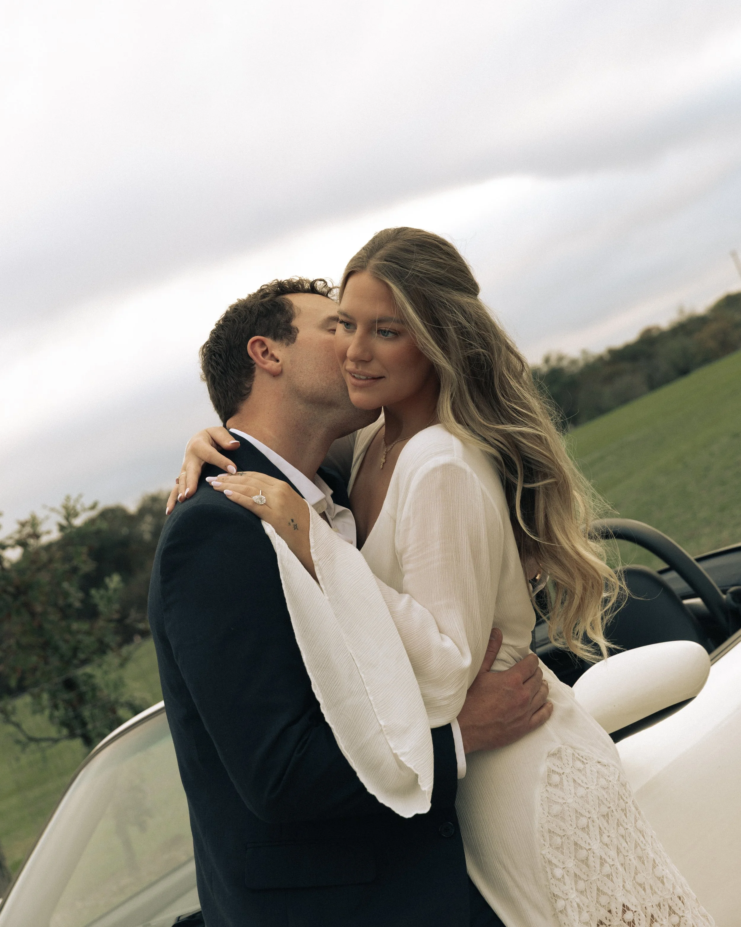 A newlywed couple embracing outdoors near a white car, with rolling green hills and a cloudy sky in the background.