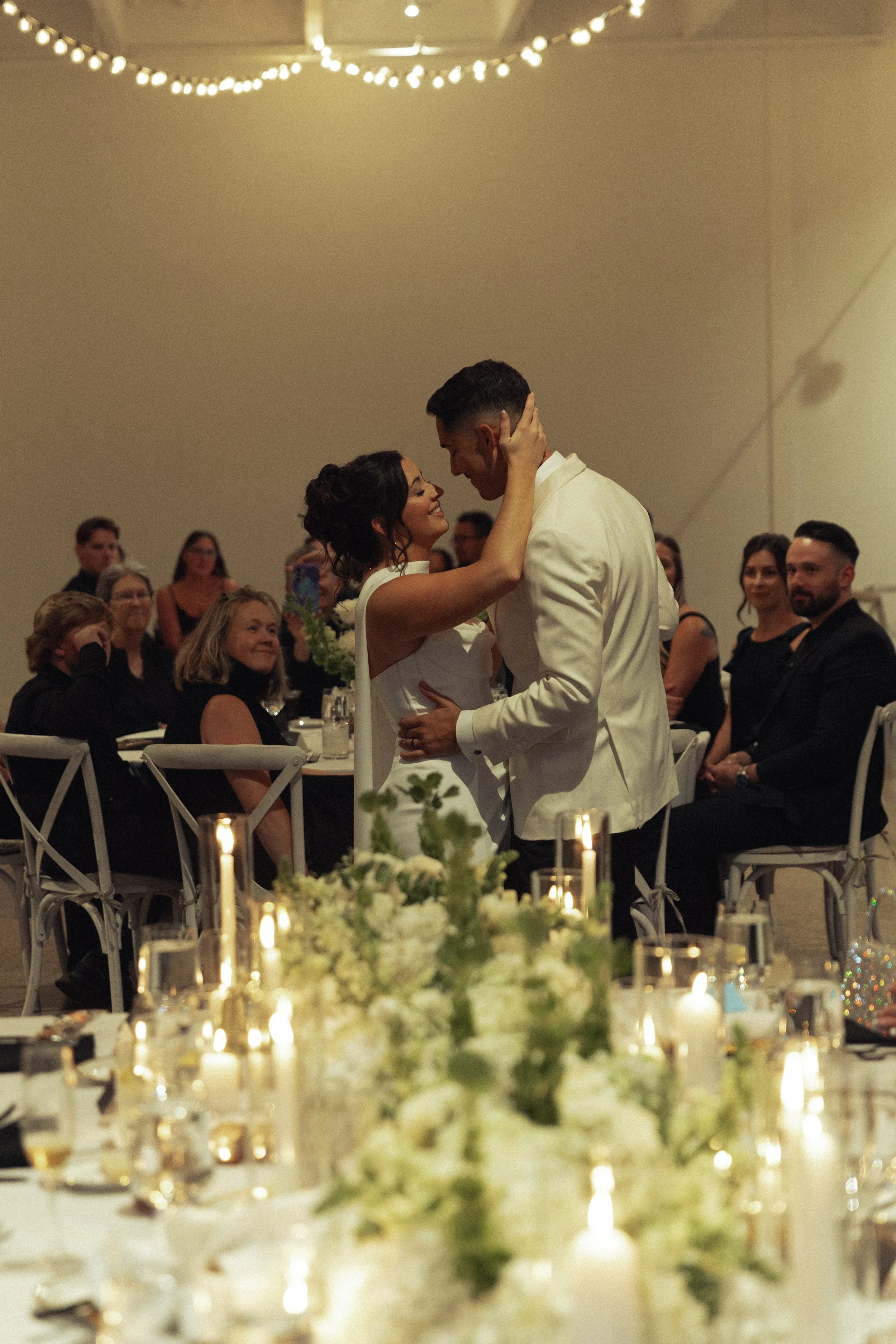 A bride and groom share a special dance at their wedding reception, surrounded by seated guests and elegant table decorations with candles and white flowers.