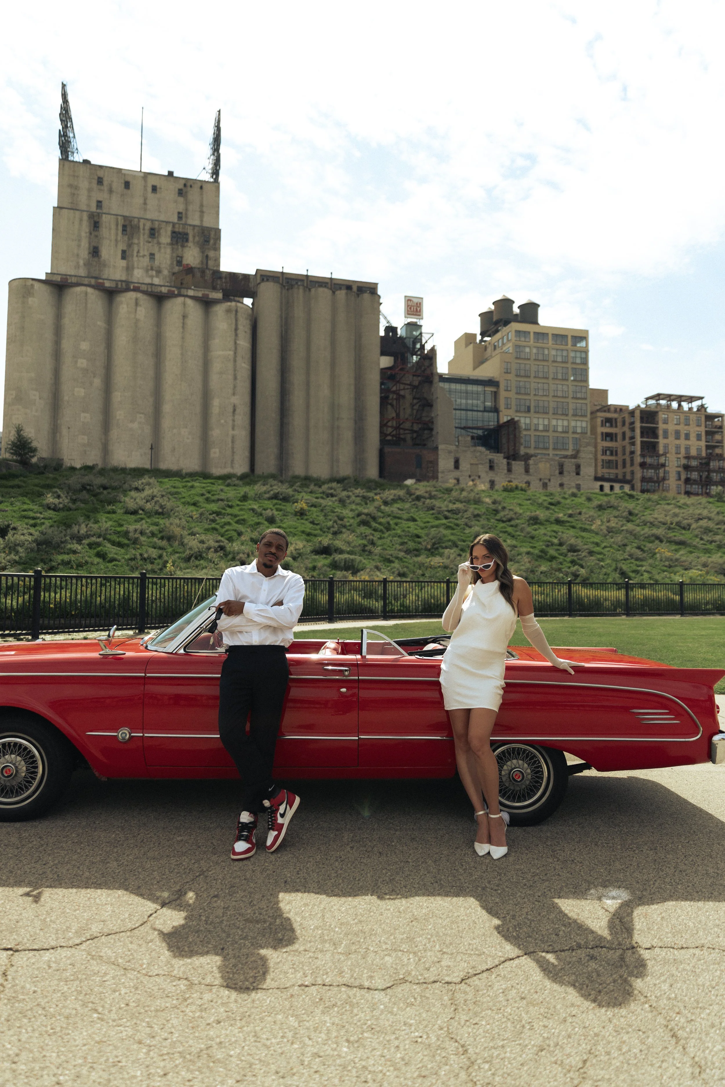 A man and a woman posed in front of a vintage red convertible car with a cityscape backdrop.