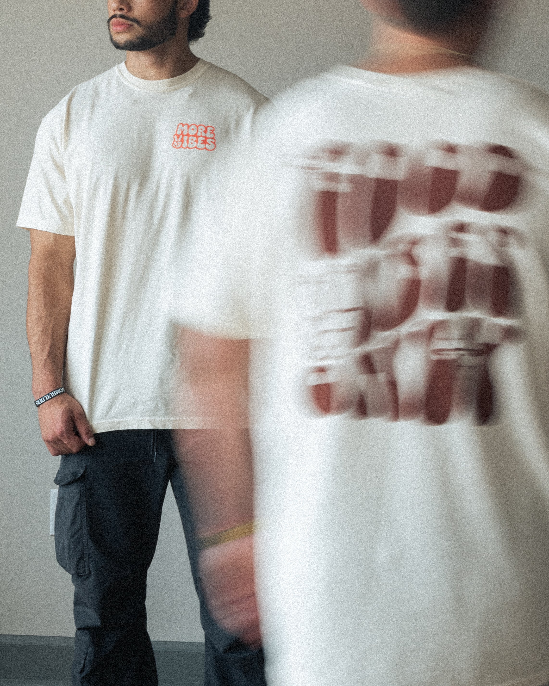 Two men wearing white T-shirts, one with red text saying 'MORE VIBES,' the other with large red text on the back; one man stands in the background, the other is mostly out of view and blurred in the foreground.