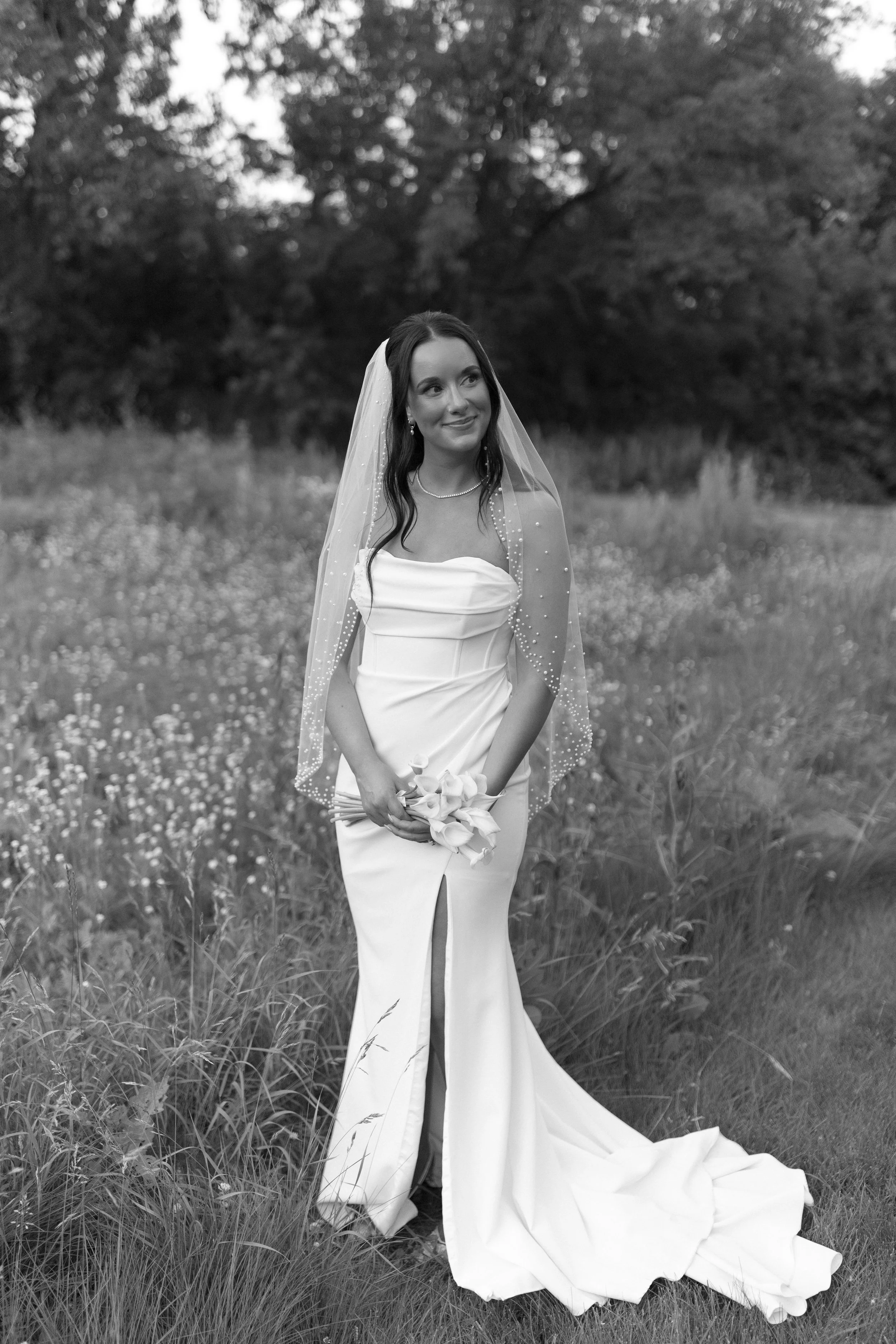 A bride in a white wedding gown holding a bouquet of flowers, standing in a grassy outdoor field with trees in the background, black and white photo.