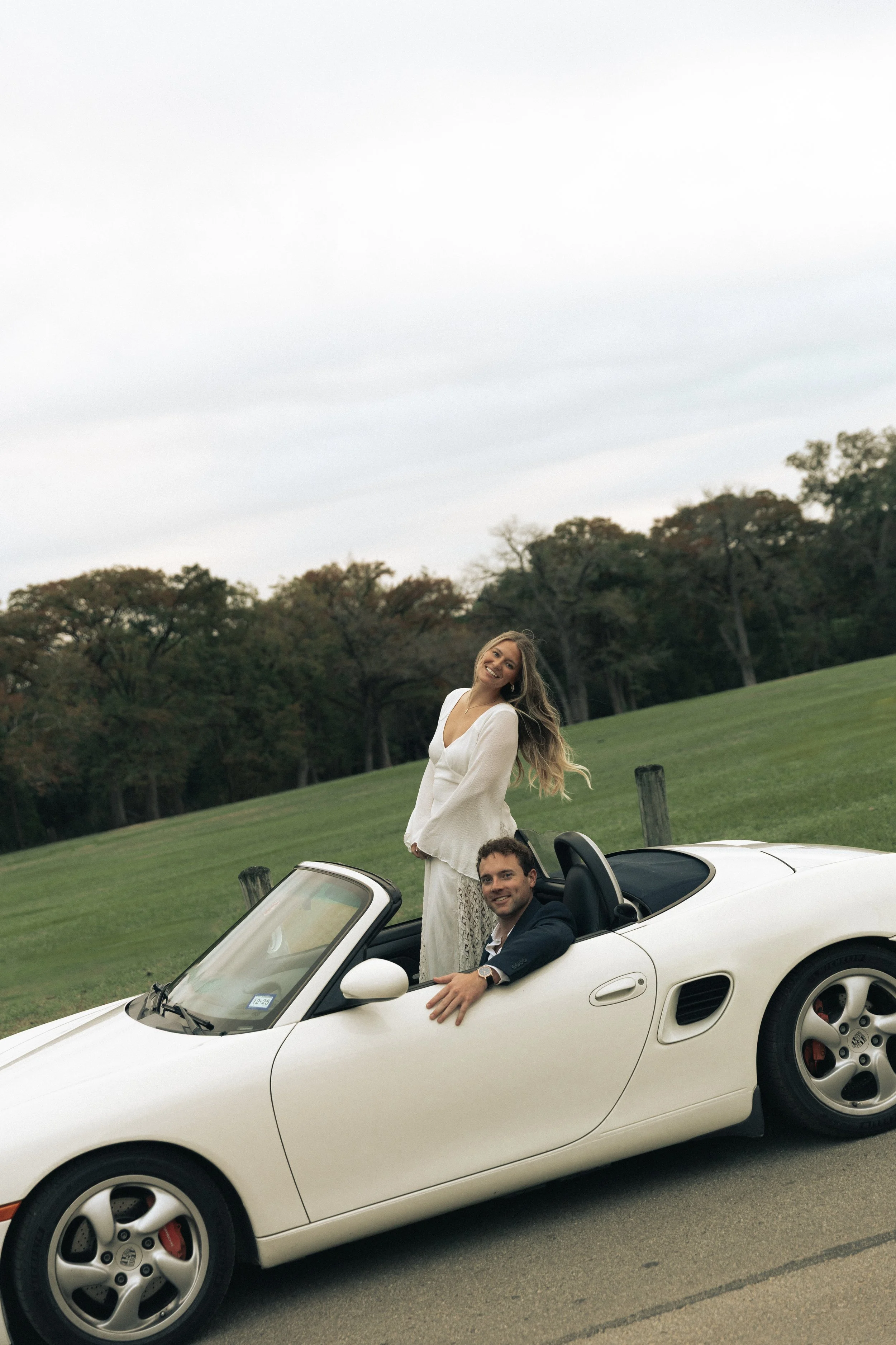 A man and woman in a white convertible car parked on a road, with trees and grass in the background, during daytime.