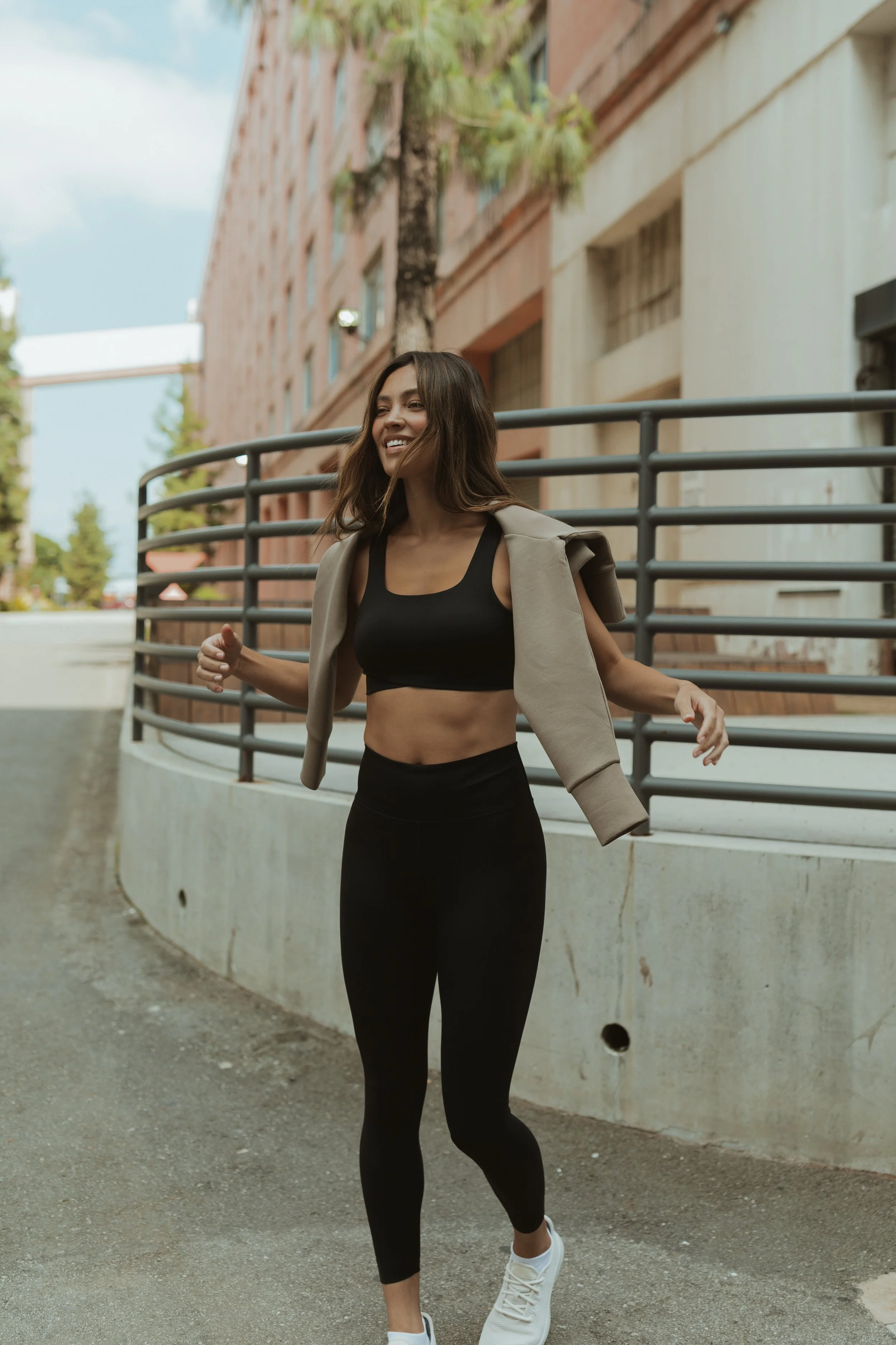 A woman jogging outdoors on a street, smiling, wearing a black sports bra, black leggings, white sneakers, and a light jacket draped over her shoulders.