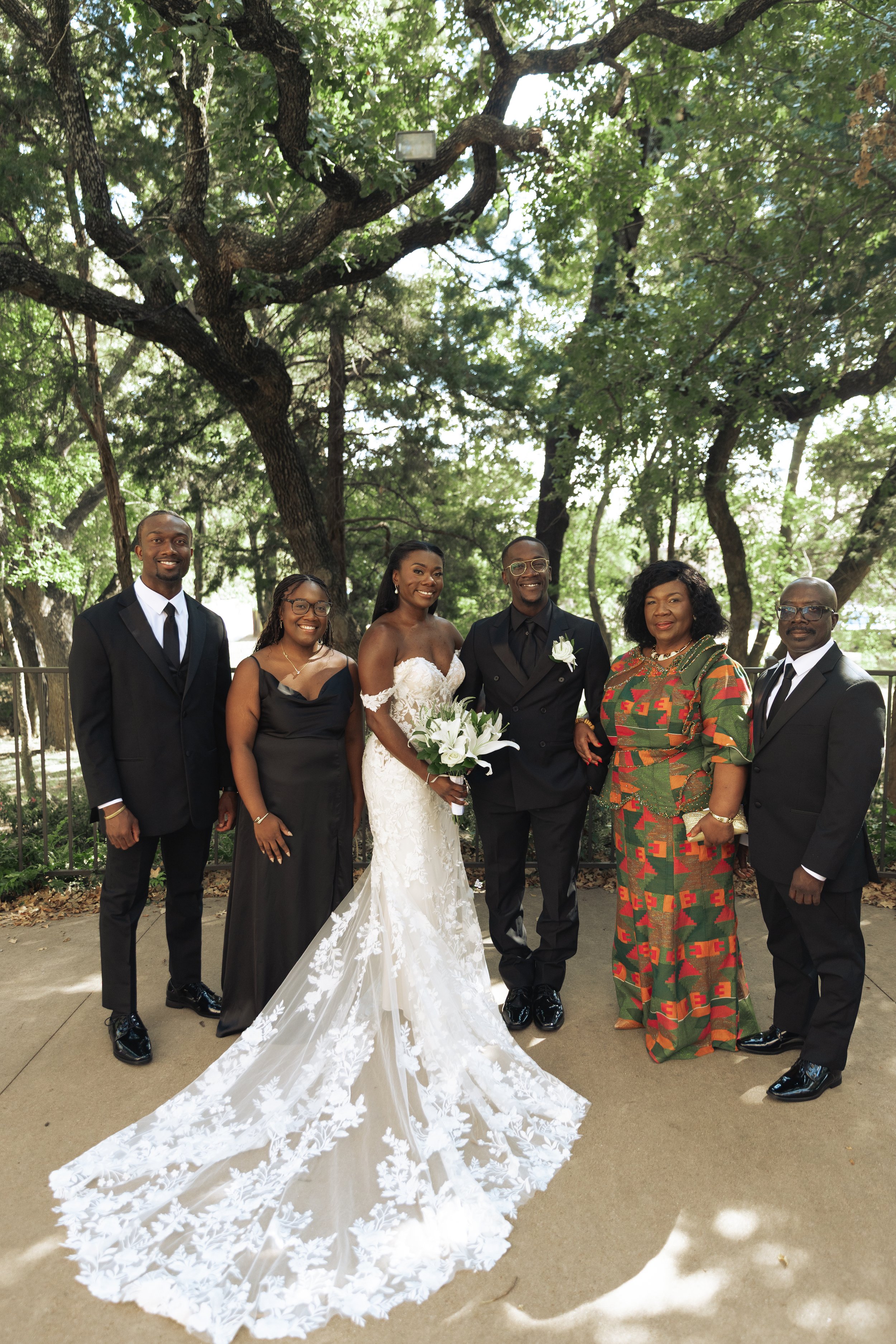 Group of six people at a wedding, standing outdoors under a large tree. The bride and groom are in the center, with the bride in a white lace wedding gown holding a bouquet, and the groom in a black tuxedo. They are surrounded by family and friends d