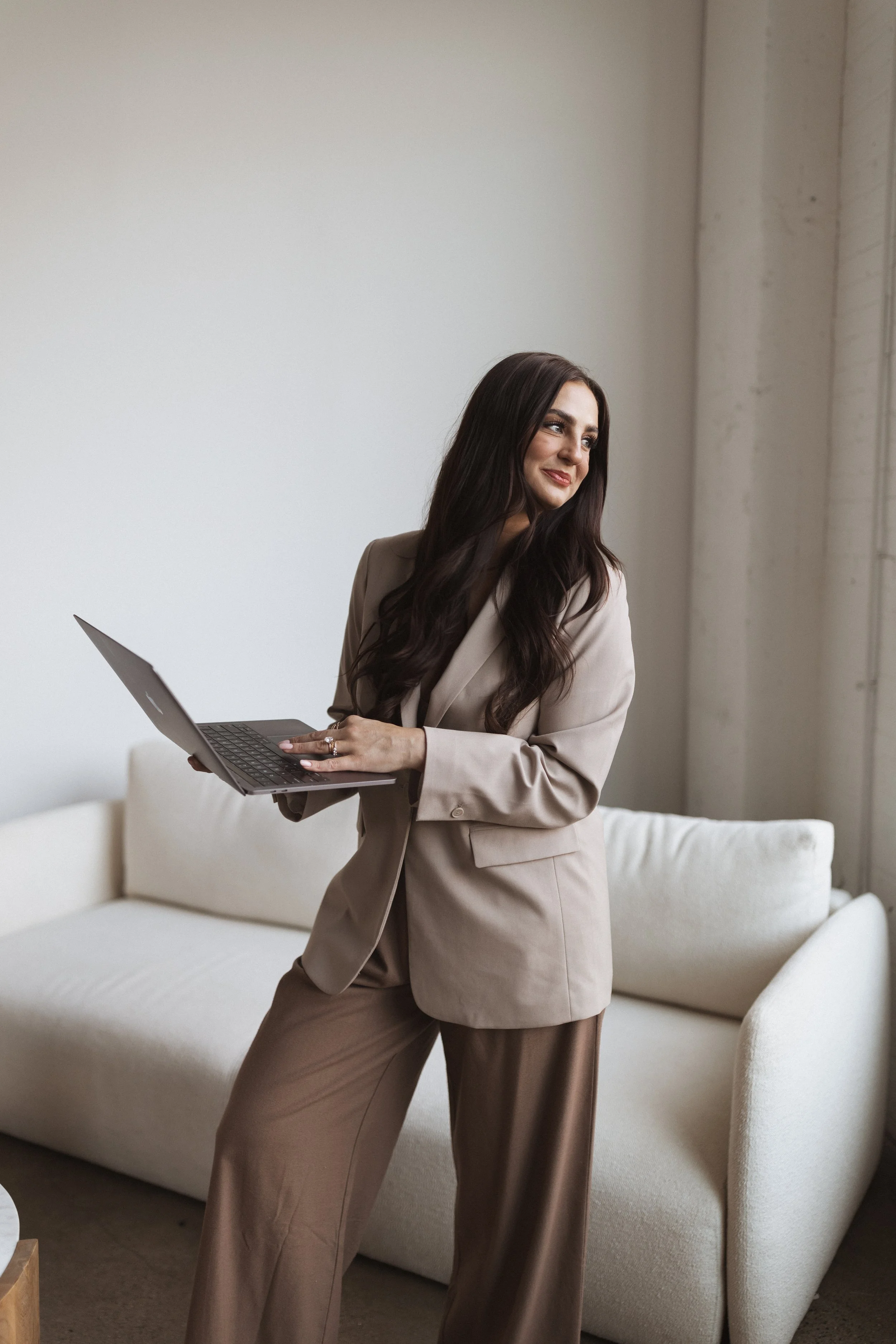 A woman with long dark hair wearing a beige suit holding a laptop and smiling, standing in a modern room with a white sofa and large window.