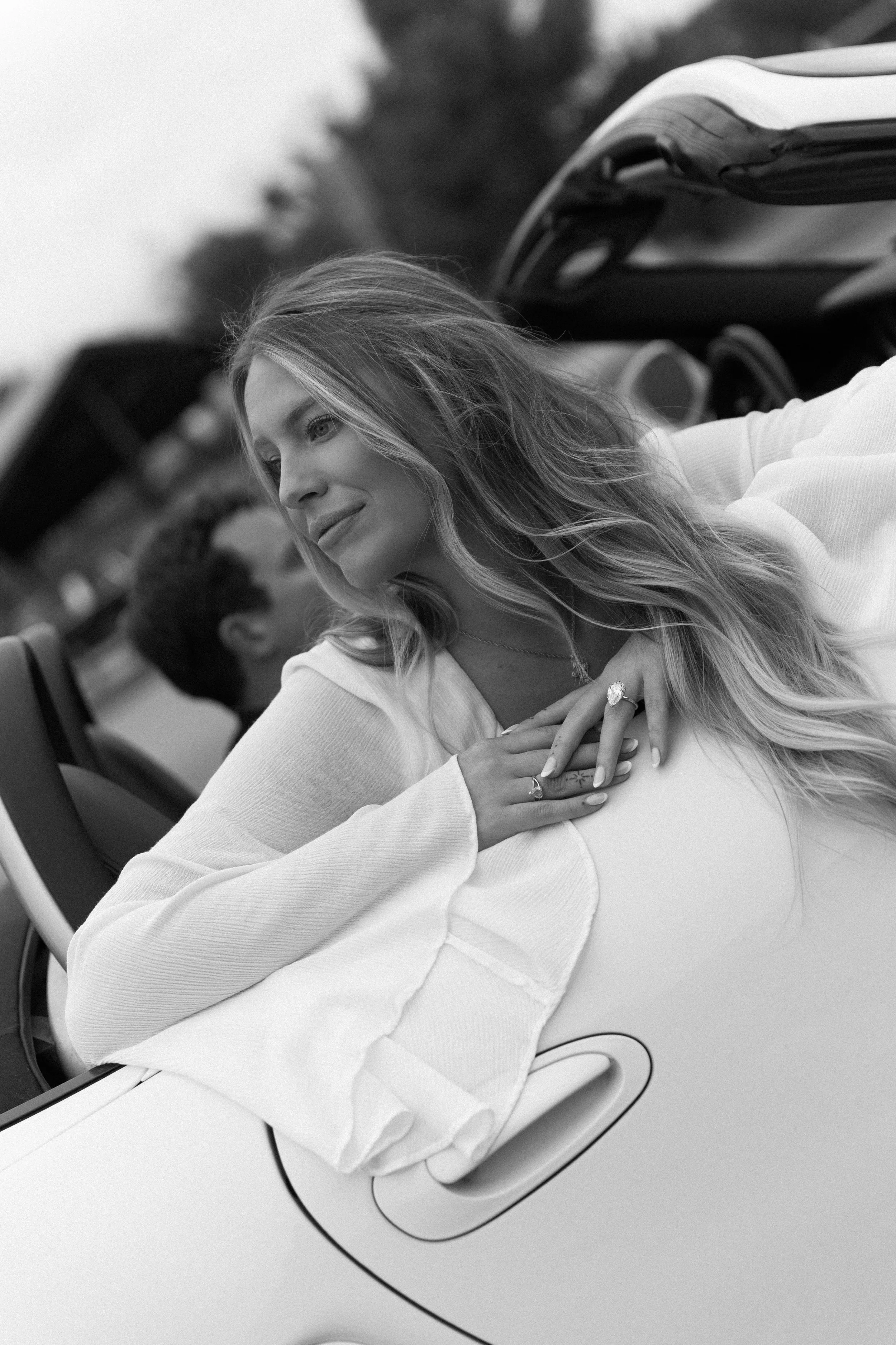 A woman with long wavy hair leaning out of a convertible car, smiling gently and looking to the side, with her hands resting on her chest and wearing rings.