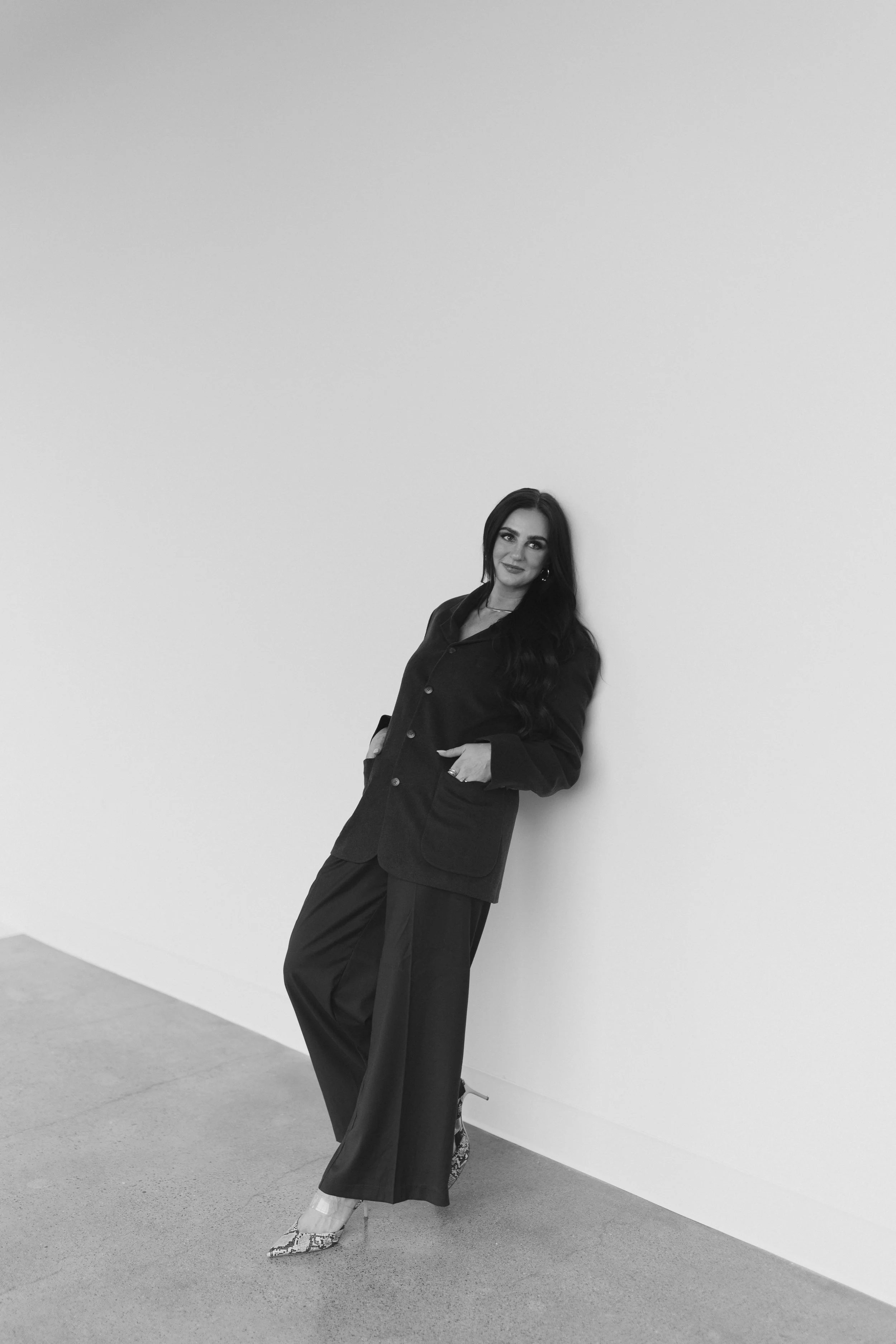 A woman with long dark hair wearing a dark blazer and wide-leg pants, standing against a plain wall, smiling slightly and posing with one hand in her pocket.